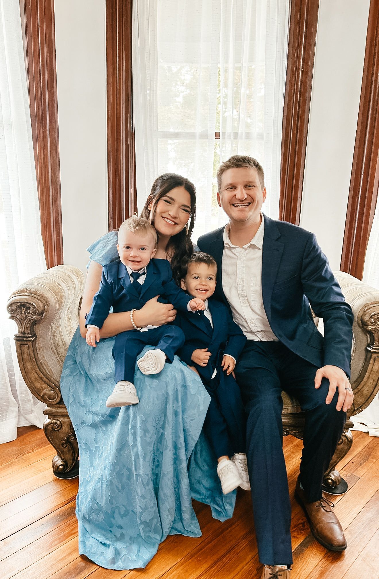 Family of four posing on ornate couch by a window; woman in blue dress, man in blue blazer, two young boys in matching outfits.