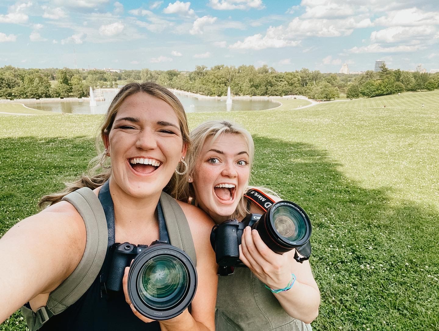 Two smiling people holding cameras outdoors near a pond and greenery.