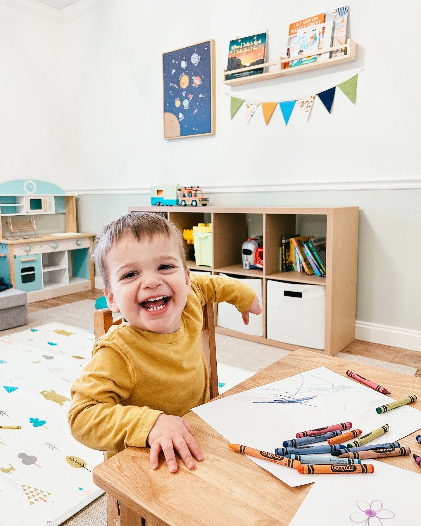 Happy child drawing with crayons at a wooden table in a playroom with a bookshelf and toys.