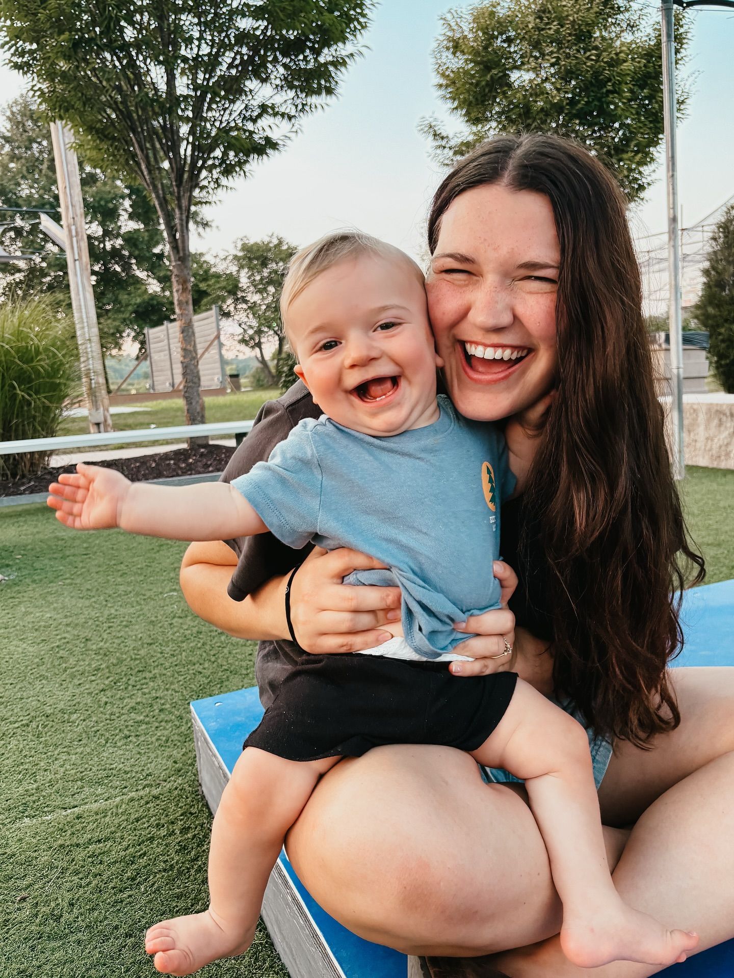 Woman holding and laughing with a baby outdoors. The baby is smiling with arms outstretched.