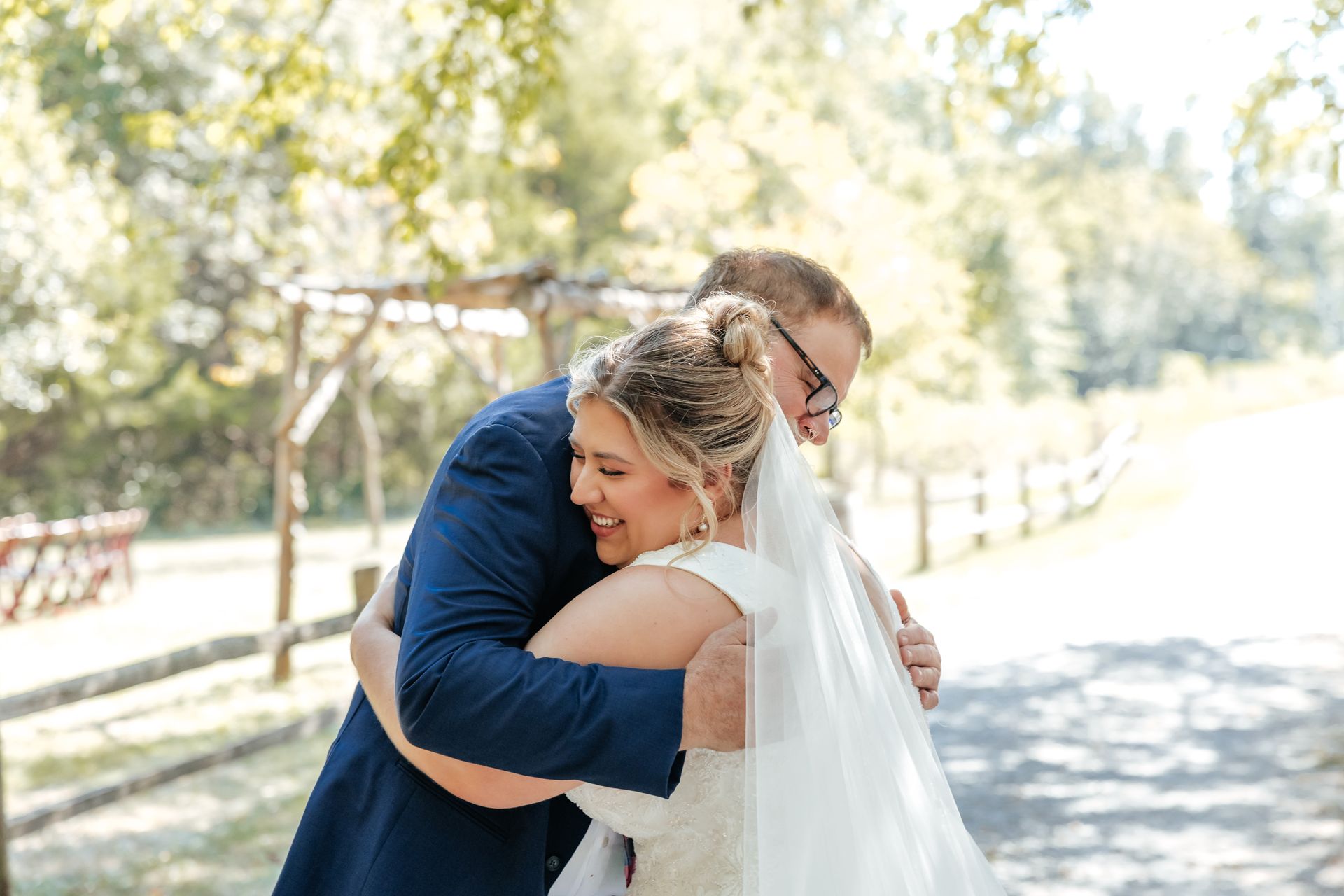 Bride and groom embrace outdoors, smiling. He wears a blue suit, she wears a wedding dress with a veil.