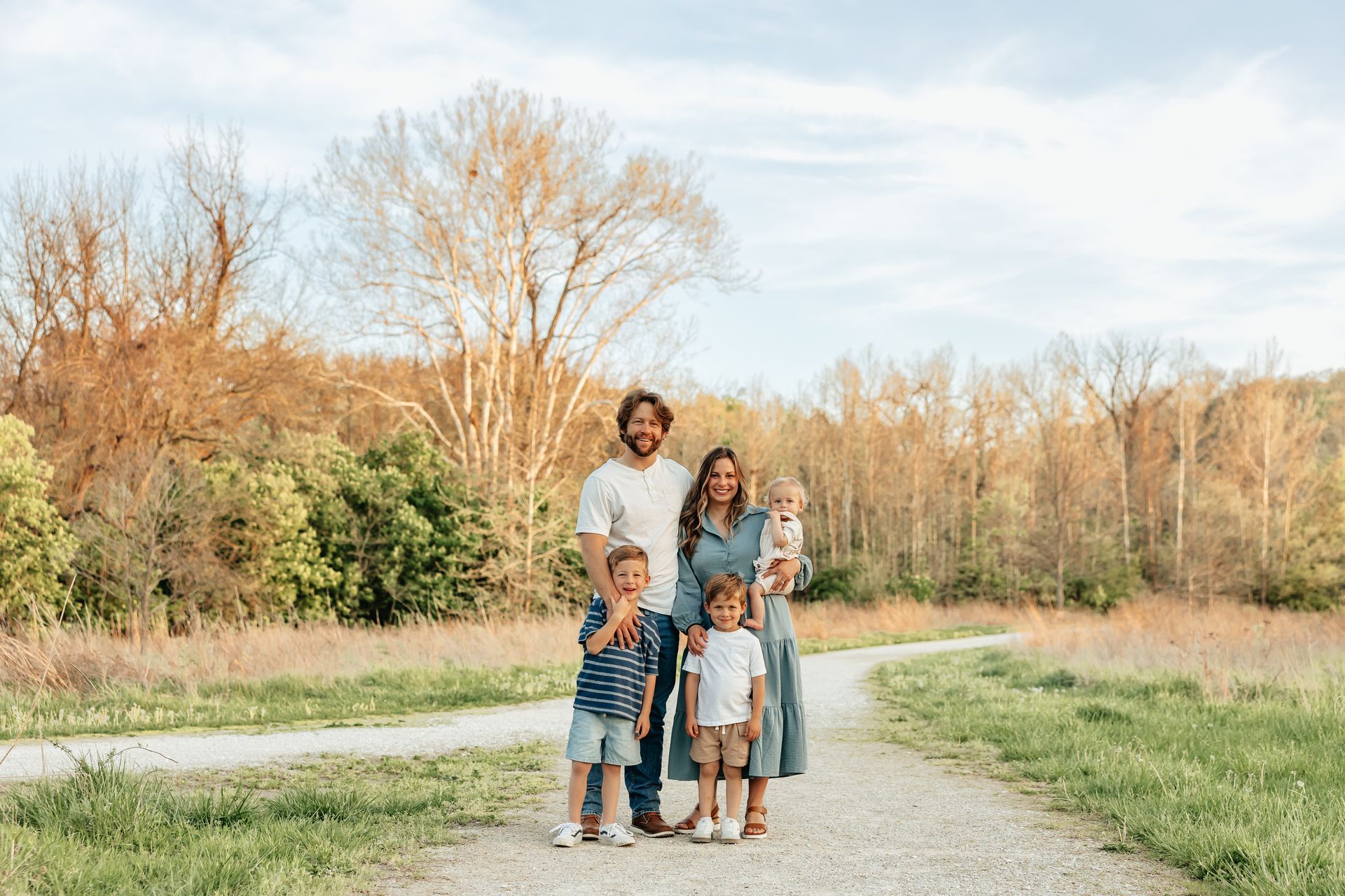 Family of five poses on a gravel path, smiling. Trees and field in background.