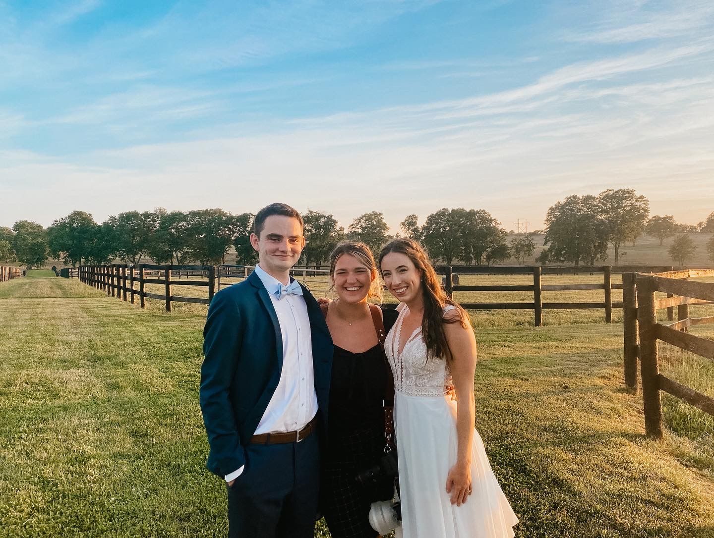 A wedding photo: Couple and a woman smiling in a field with a wooden fence. Sun setting in the distance.