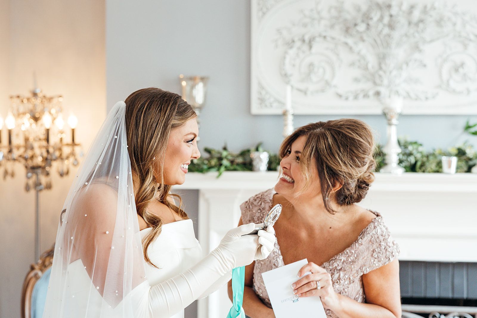 Bride and mother laugh together, holding gifts. Bride wears veil and gloves, mother holds card near a fireplace.