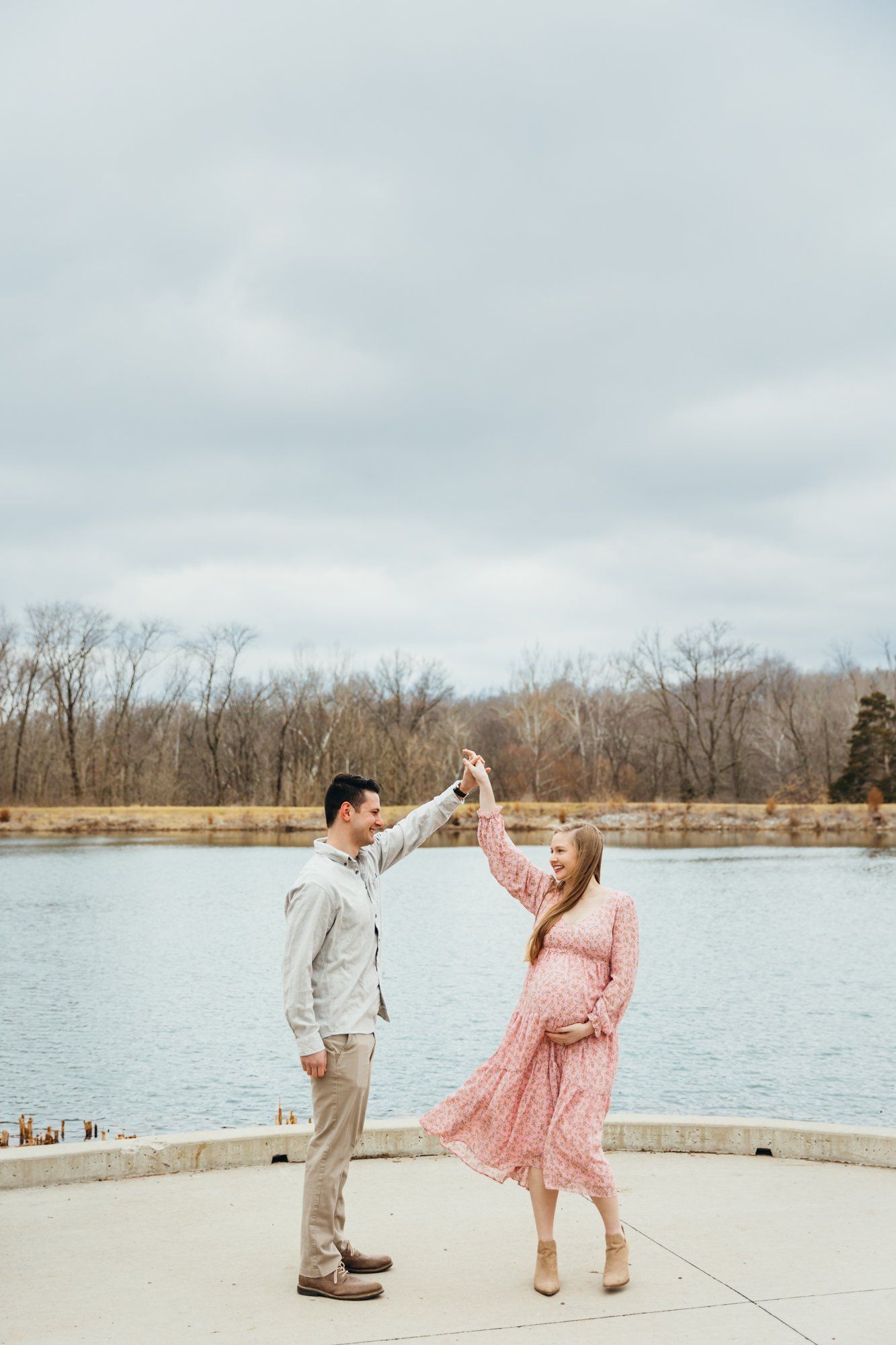 Husband spins his pregnant wife as they dance in front of a lake