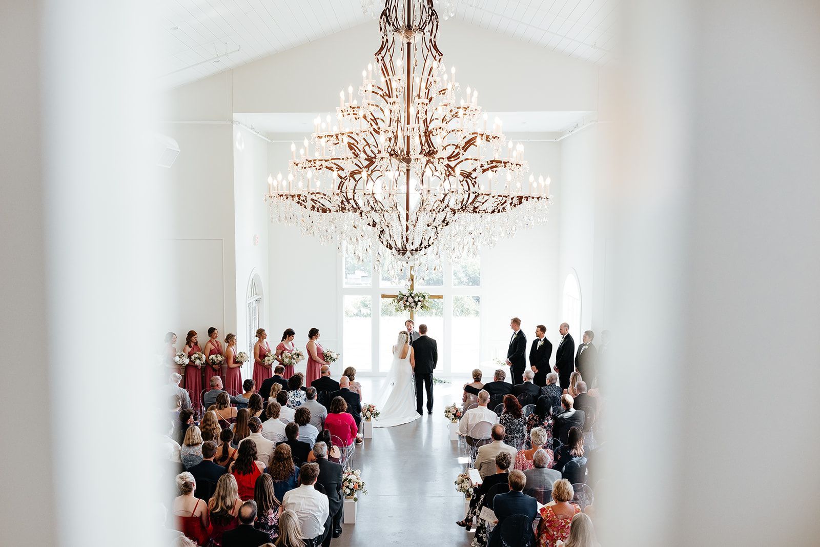 Wedding ceremony in a bright white chapel with a large chandelier; bride and groom at altar.
