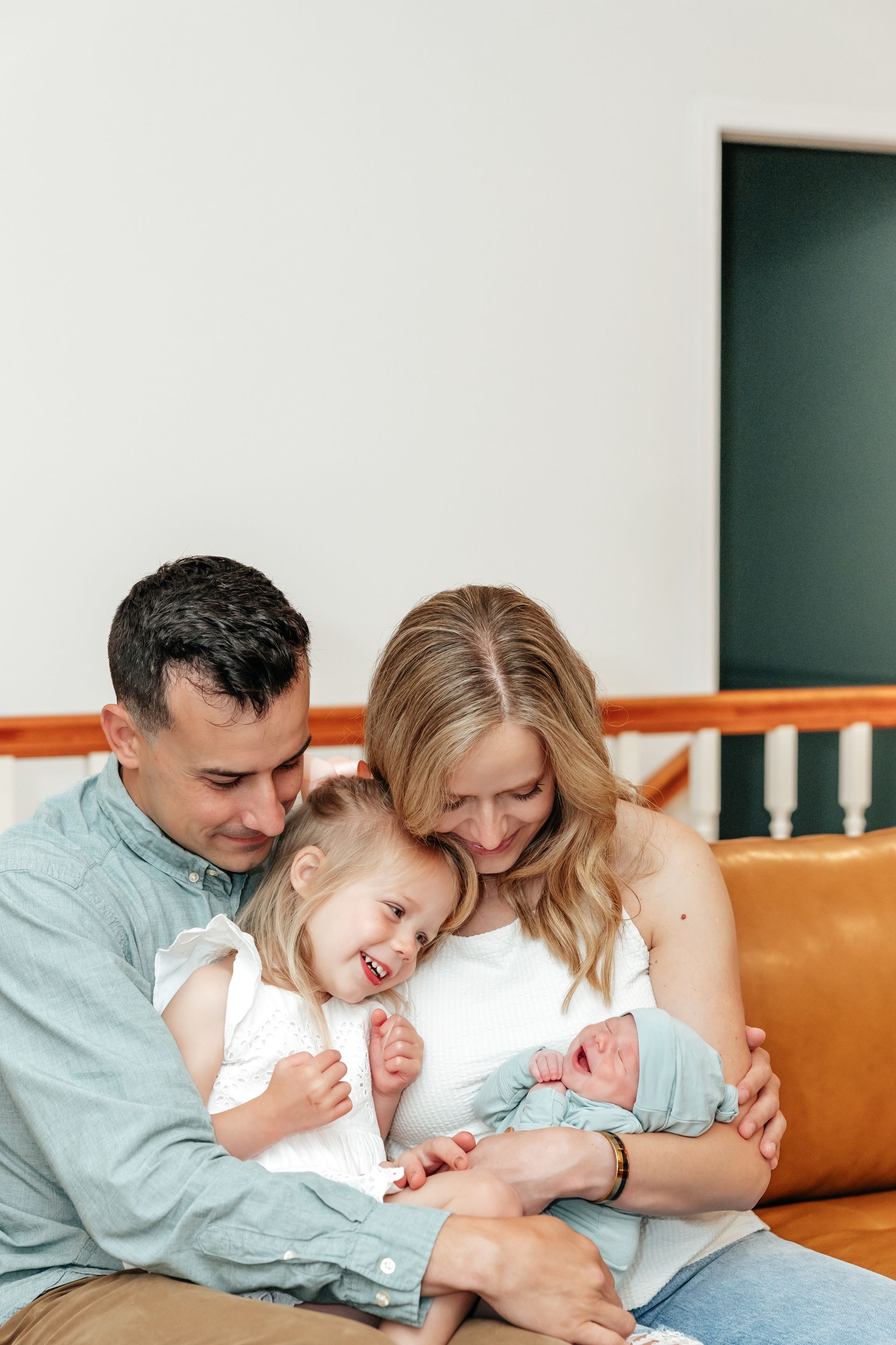 Family of four sitting on a couch. The parents are smiling at their two children; one is a newborn.