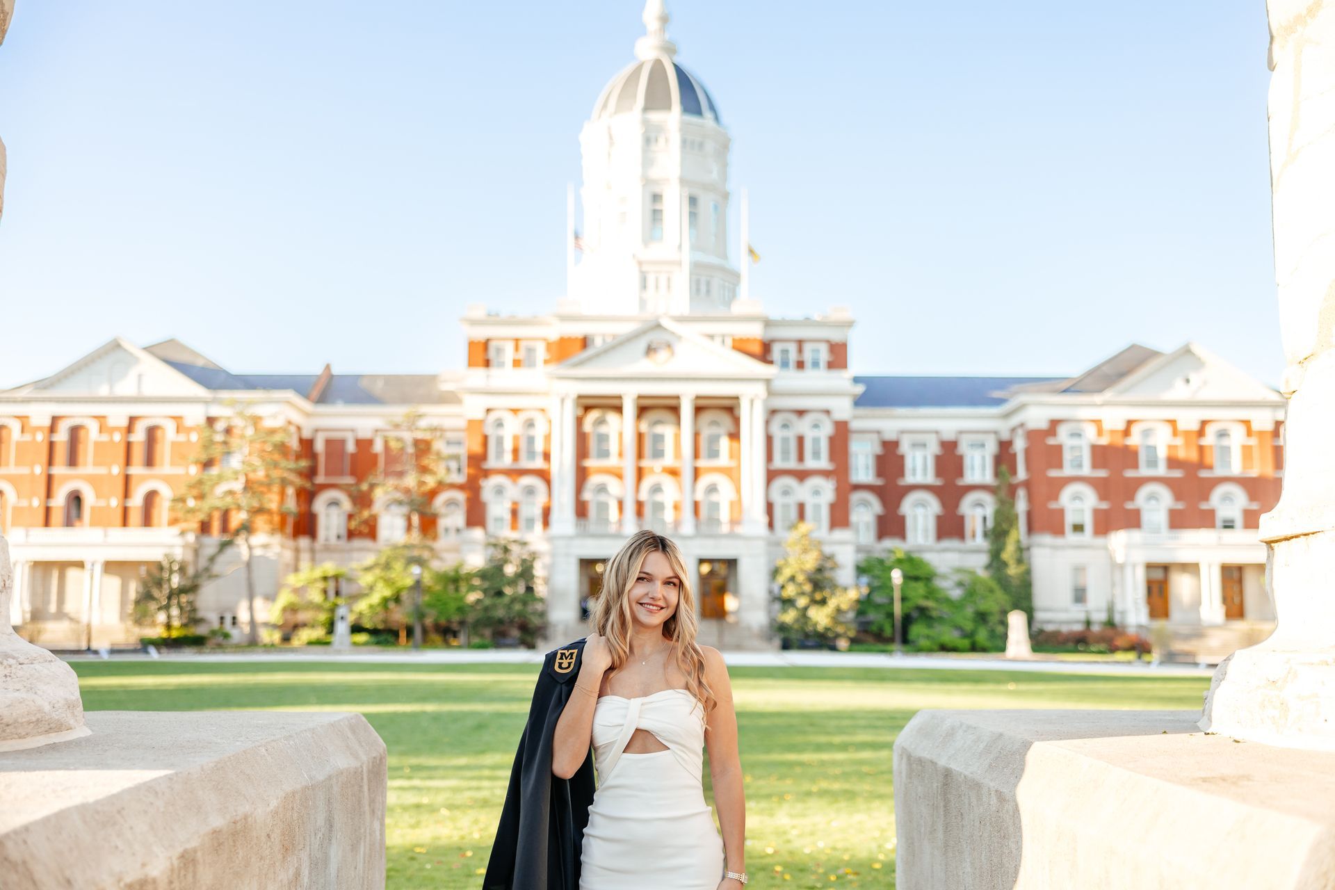 Woman in white dress smiles, poses in front of a large brick building with a dome. Black jacket over shoulder.