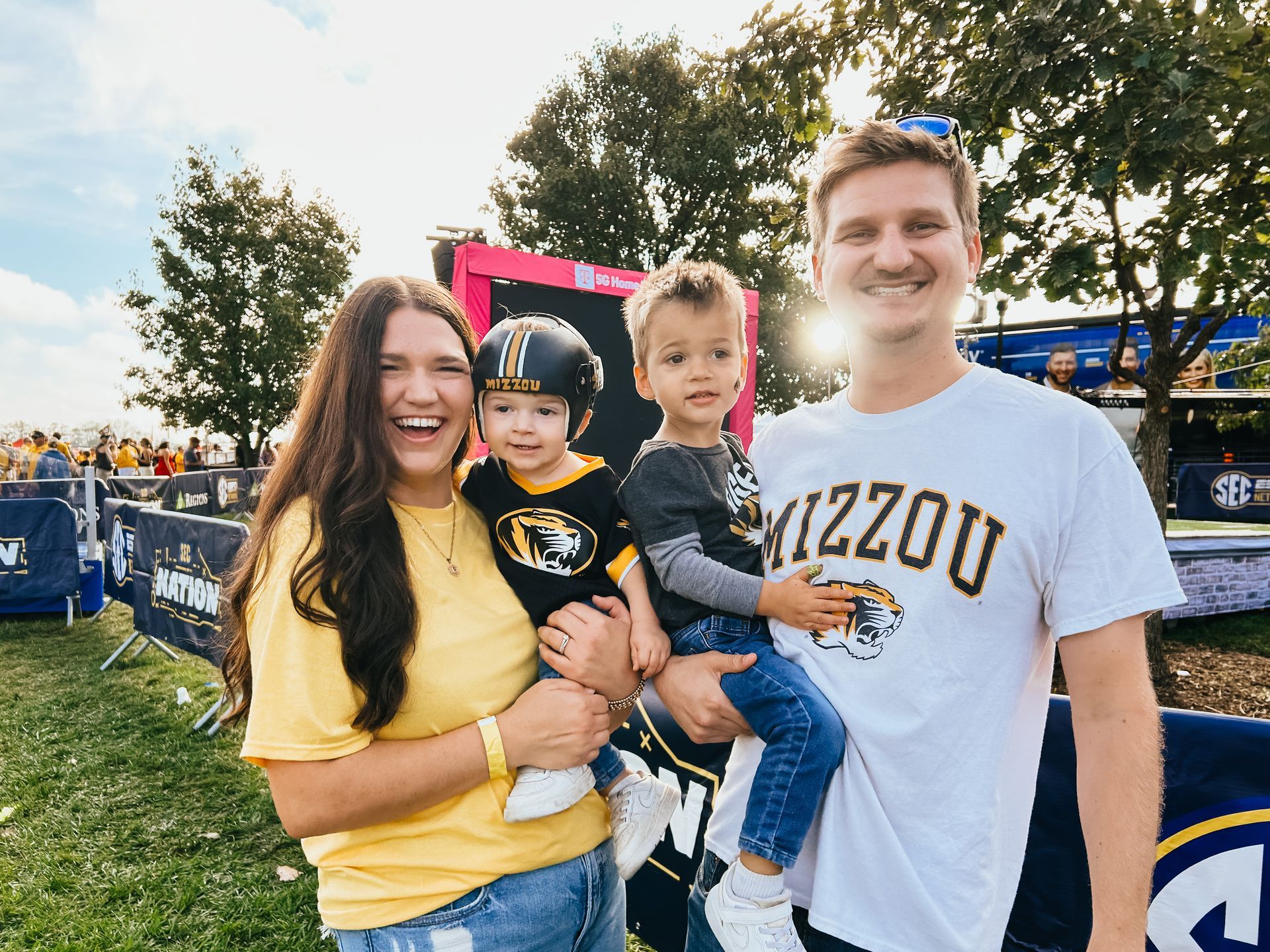 Family, woman holding toddler in helmet, man holding another child, both wearing Mizzou gear, smiling outside.