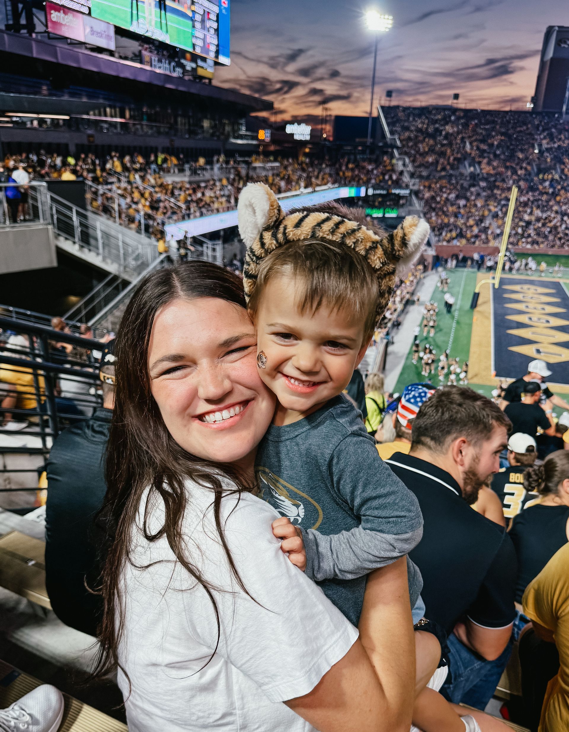 Woman holds a child wearing tiger ears at a football stadium. Both smile. Evening sky and crowd visible.