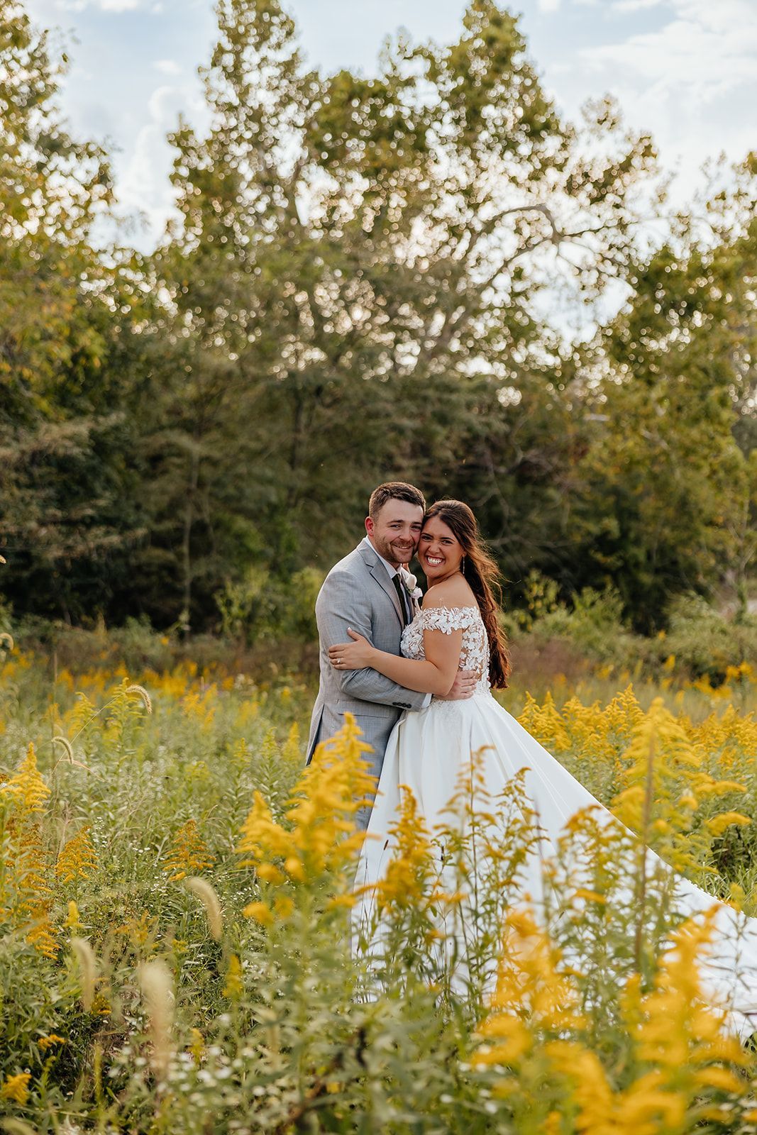 Couple embracing in a field of yellow flowers, dressed in wedding attire, with a backdrop of trees.