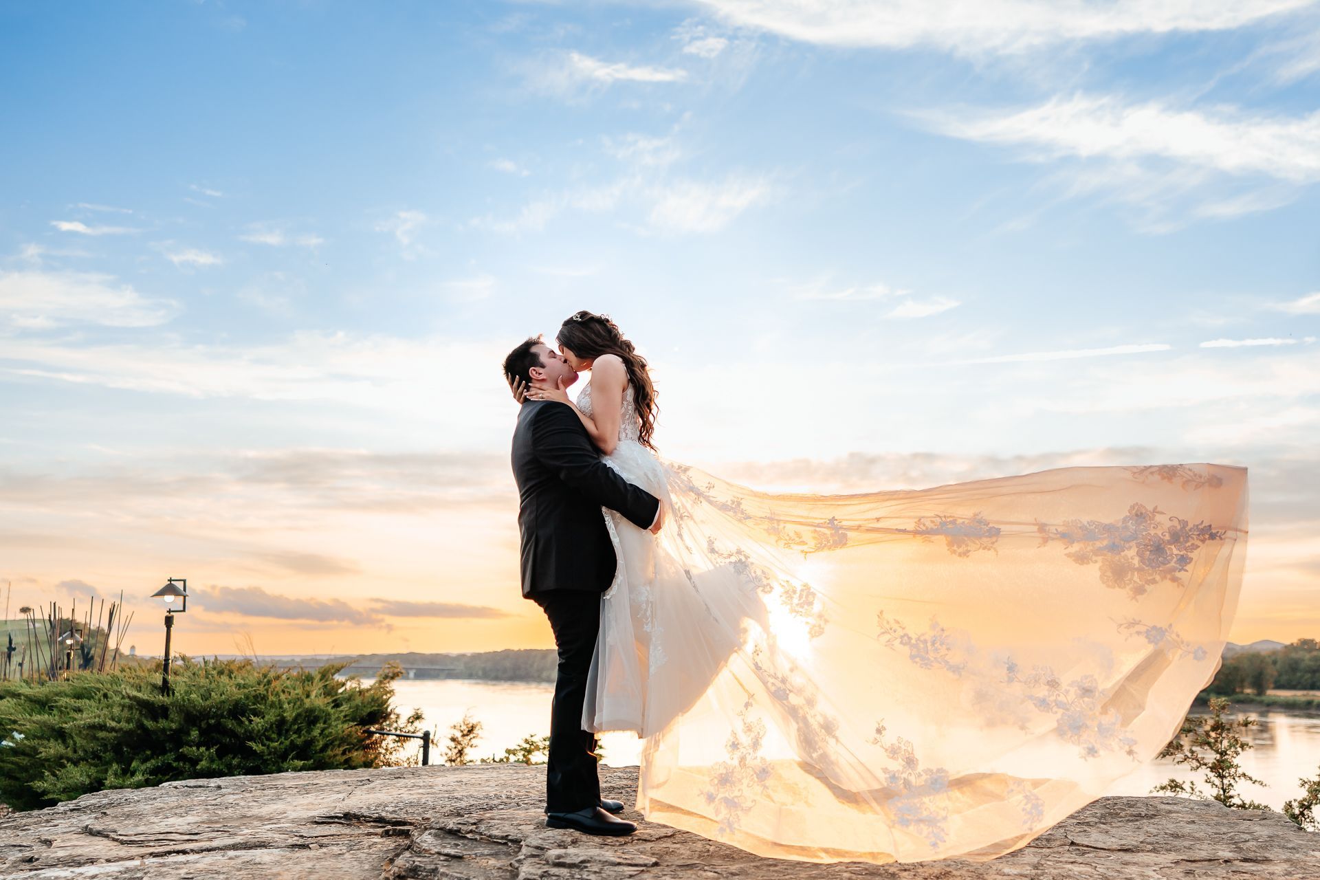 Groom lifting bride, their faces close, dress flowing in the wind against a sunset backdrop.