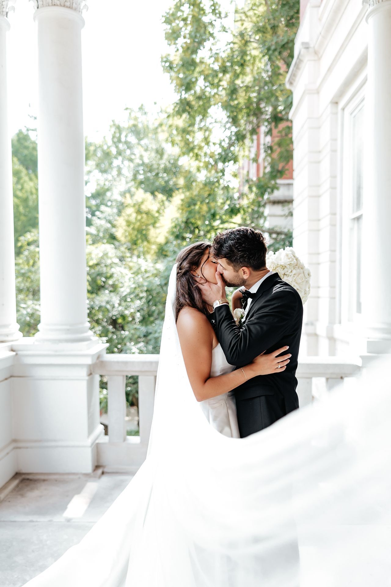 Couple kissing on a white porch, woman in wedding dress, man in a tuxedo, sunlight.