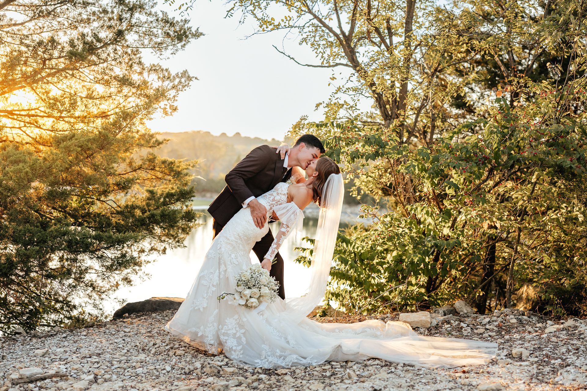 Couple kissing in a dip, bride in a white lace dress with a long train, groom in a black suit, with trees and water in the background.