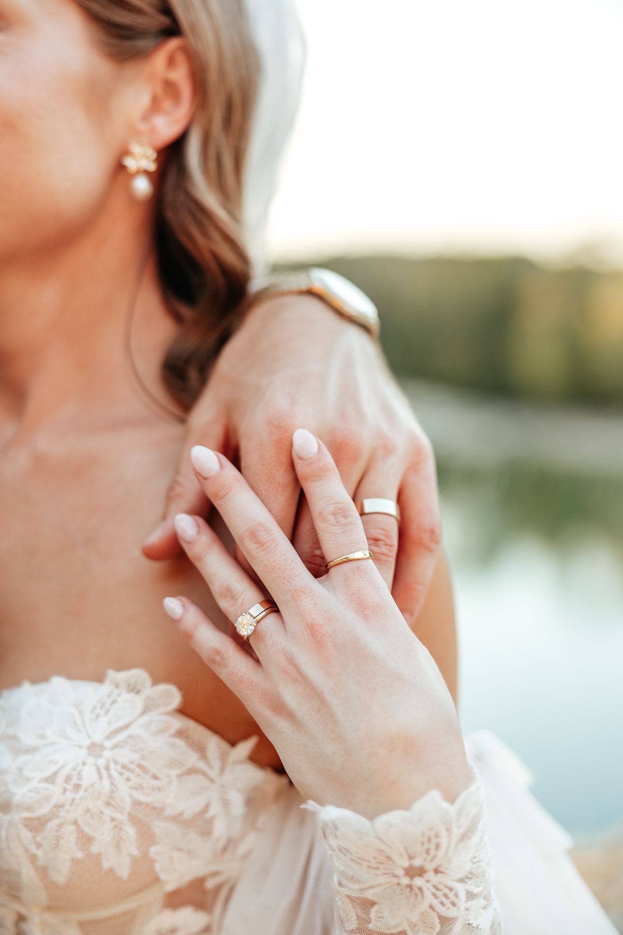Bride and groom holding hands, showing off wedding rings, with a scenic lake in the background.