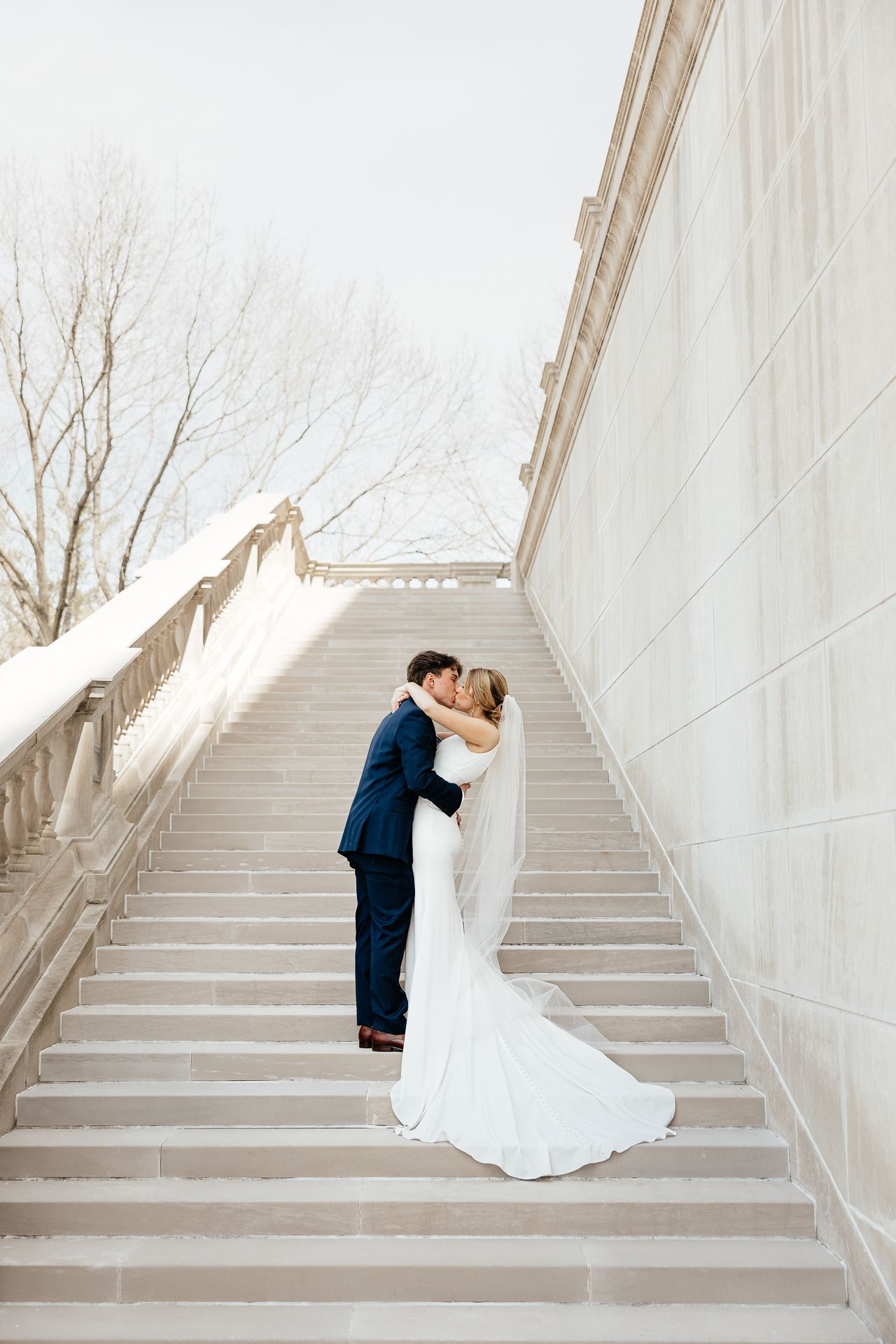 Bride and groom kissing on stone stairs; man in blue suit, woman in white gown and veil.