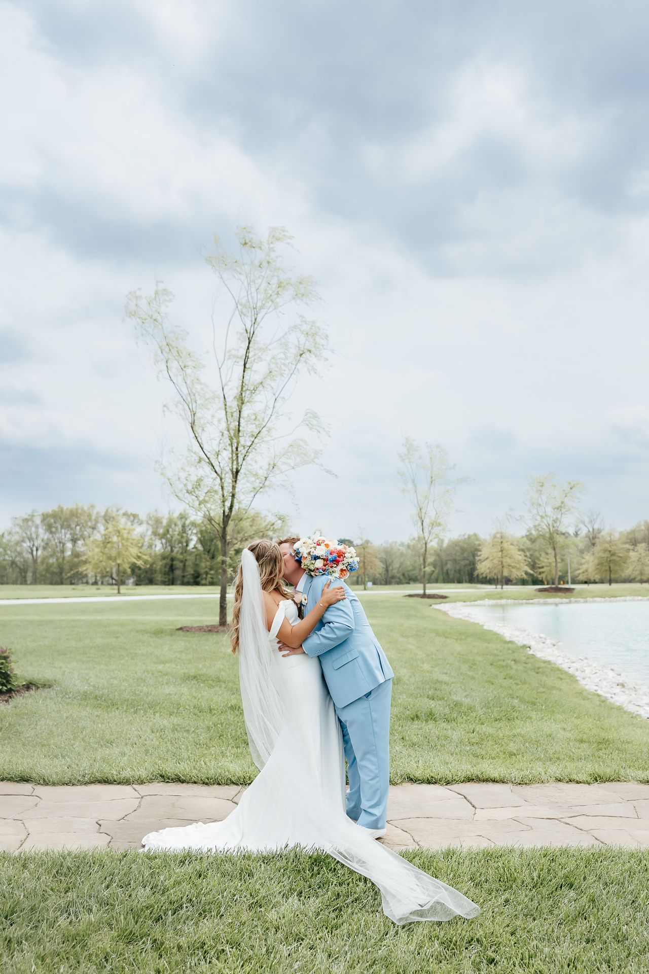 Bride and groom embrace and kiss outdoors by a pond; the bride wears a white dress and veil, the groom, a light blue suit.