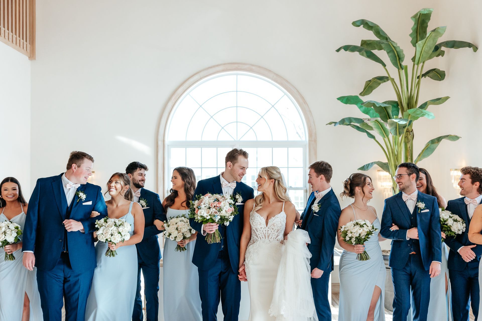 Wedding party poses in a bright room with the bride and groom looking at each other, flanked by bridesmaids in blue and groomsmen.