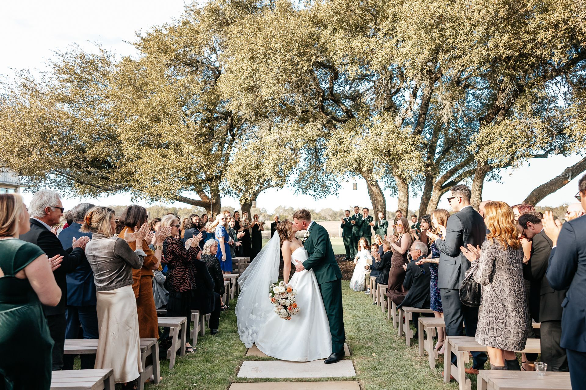 Newlyweds kiss as guests clap during an outdoor wedding ceremony under trees.