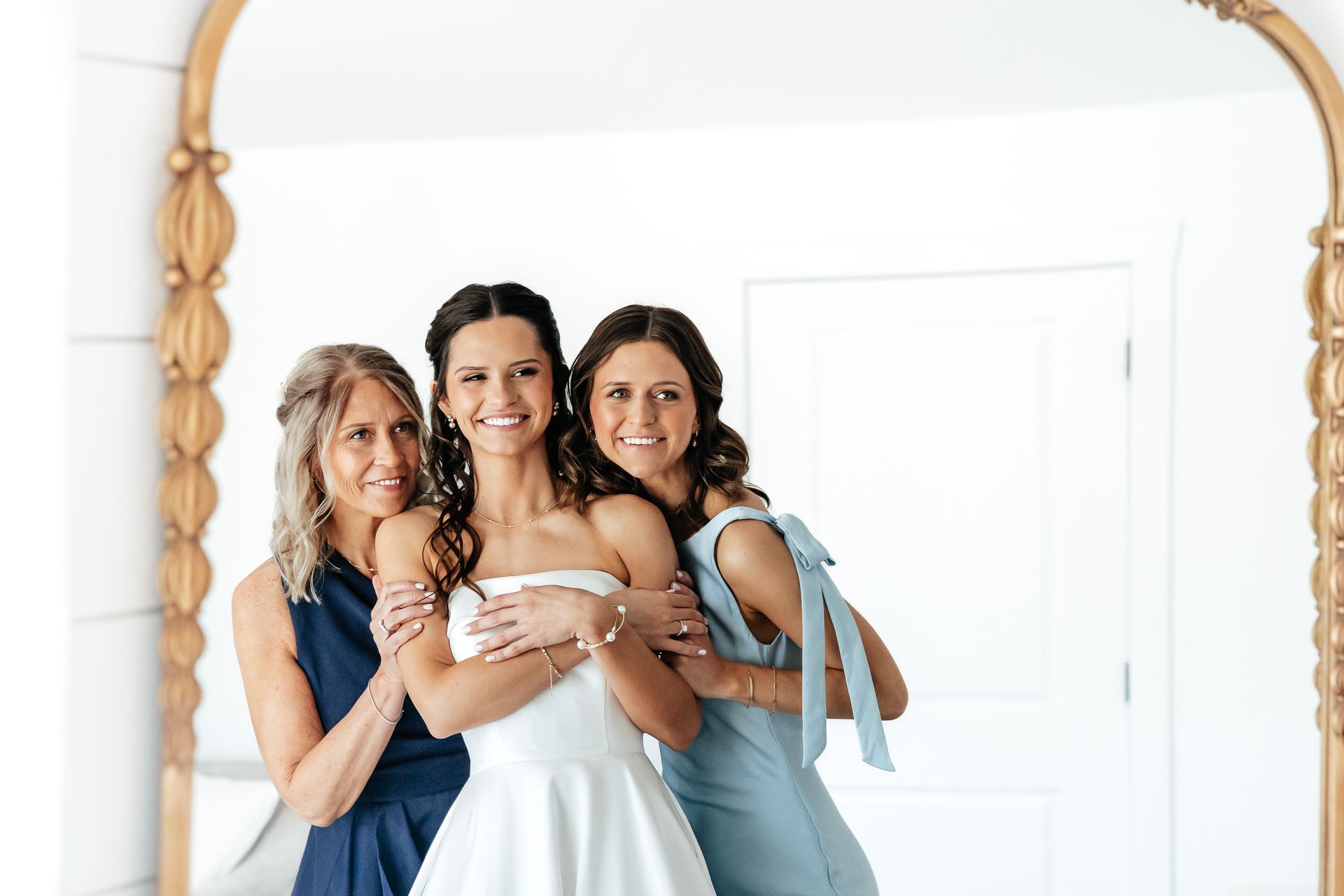 Bride with two women smiling in front of a mirror; one in a blue dress and the other in a navy top.