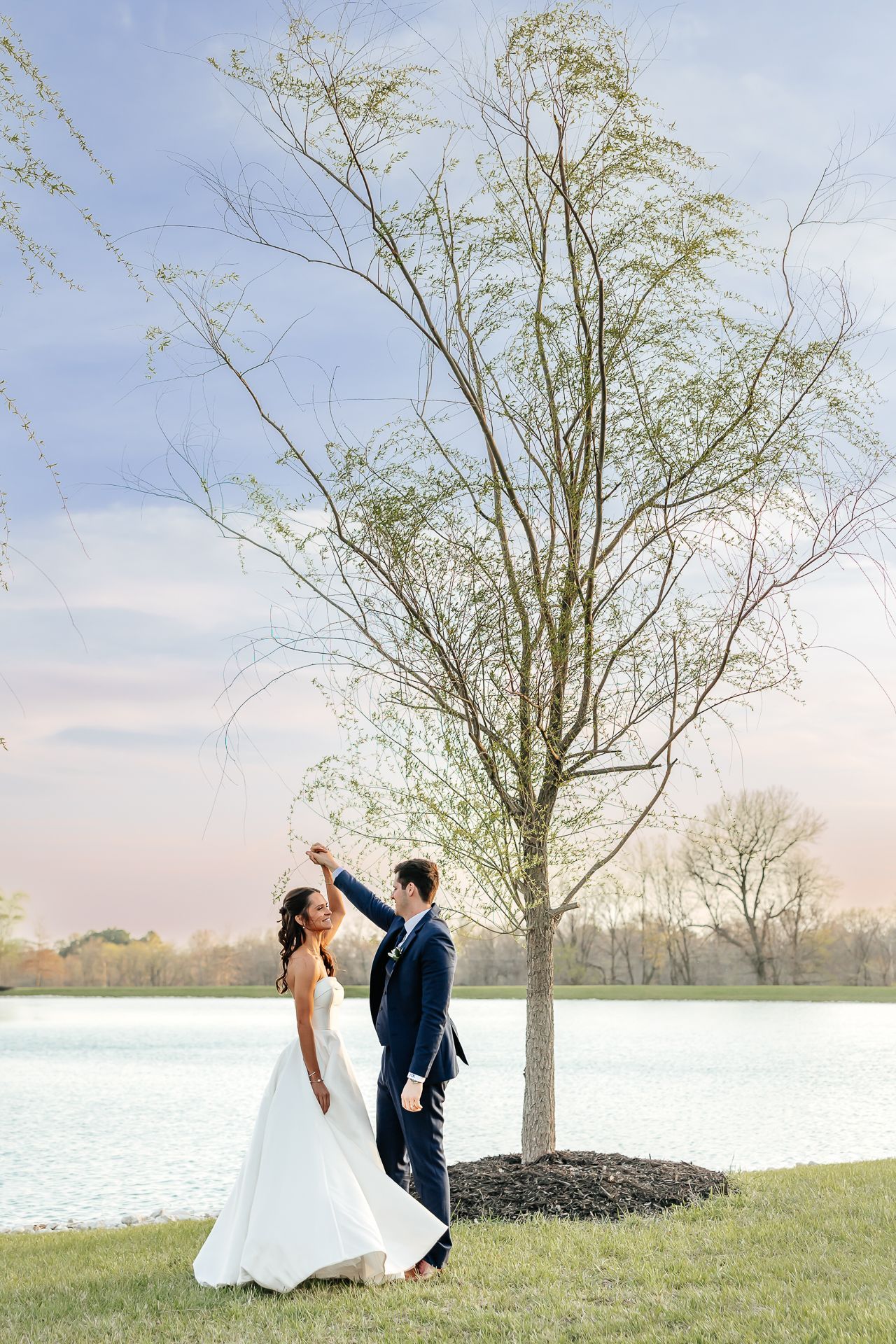 Couple dancing near a tree by a lake, the woman in a wedding dress, the man in a suit, with a blue sky.