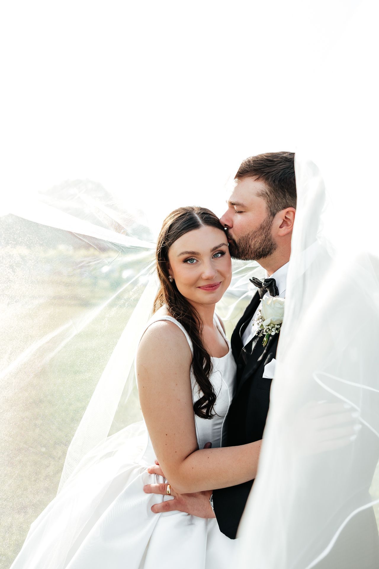 Bride and groom embracing, the groom kissing the bride's forehead. They are under a sheer veil, outside.