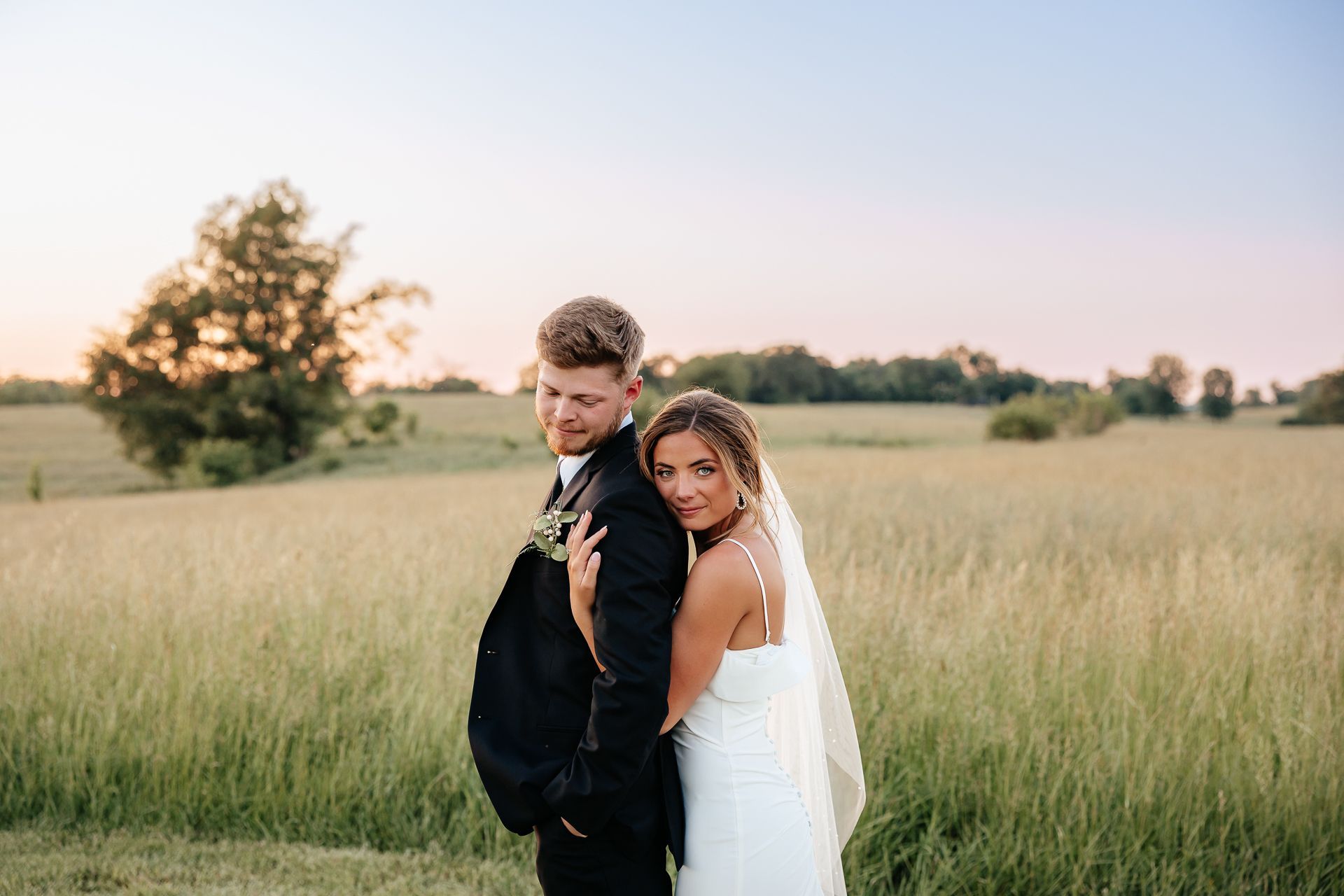 Bride and groom embrace in a field at sunset. The bride leans on the groom, both smiling.