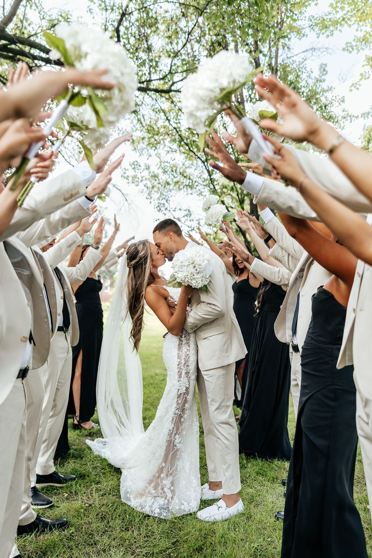 Bride and groom kiss as guests raise bouquets, forming an archway. Outdoor wedding setting with greenery.