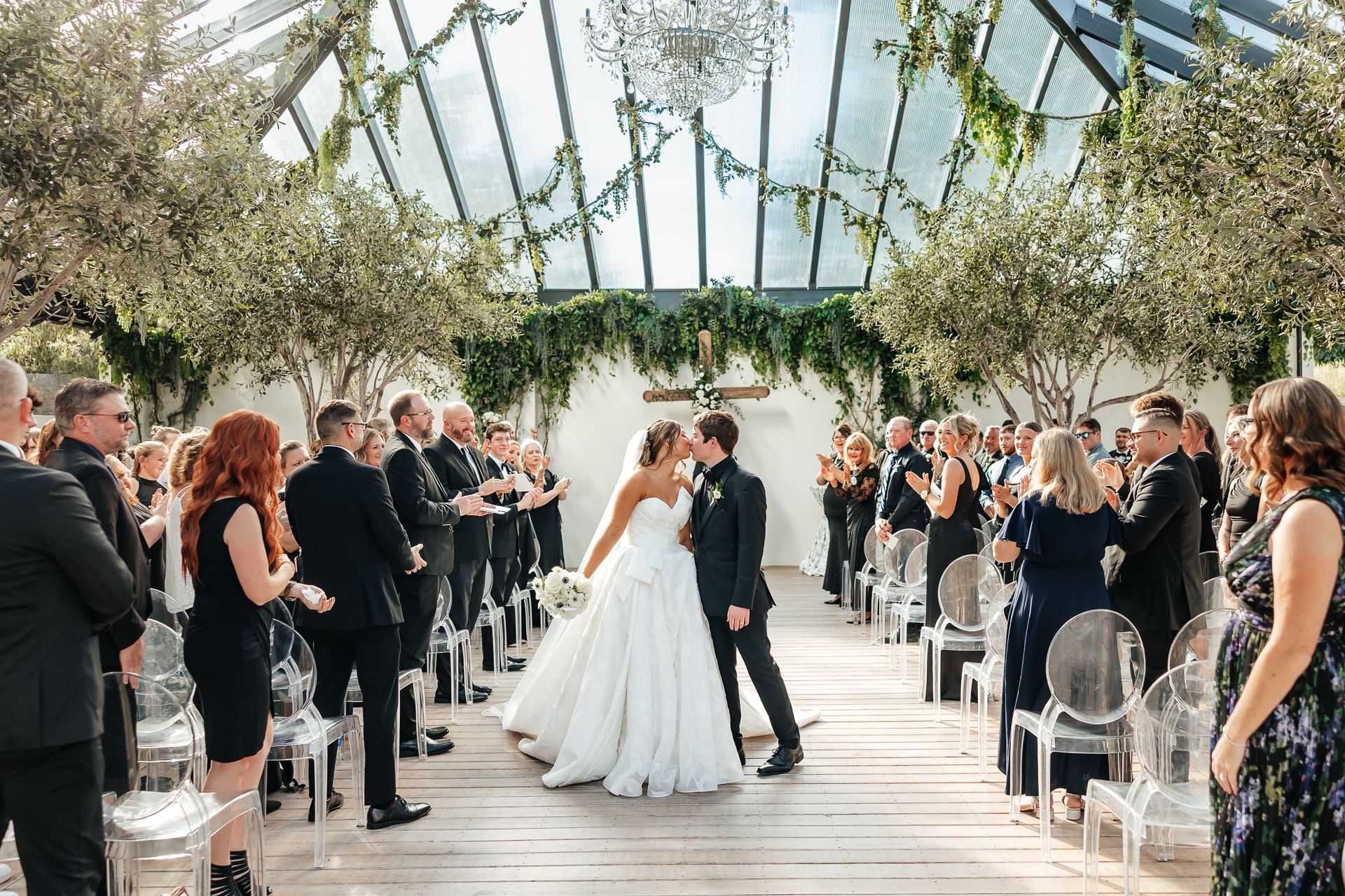 Wedding ceremony: couple kissing, guests applauding, in a glass-roofed venue with greenery and trees.
