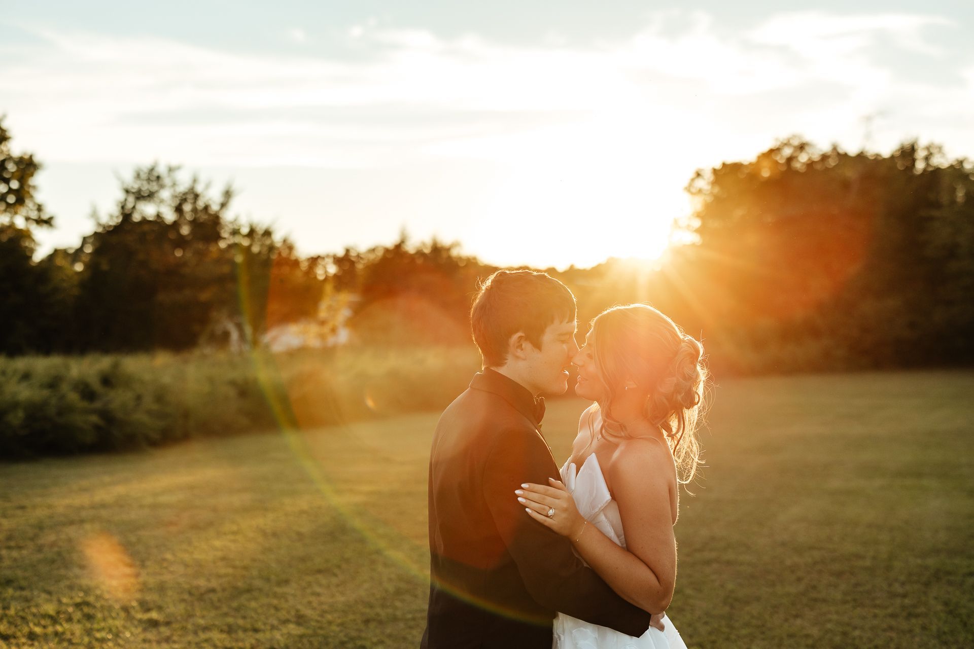 Couple embracing at sunset in a field; sun flares around them.