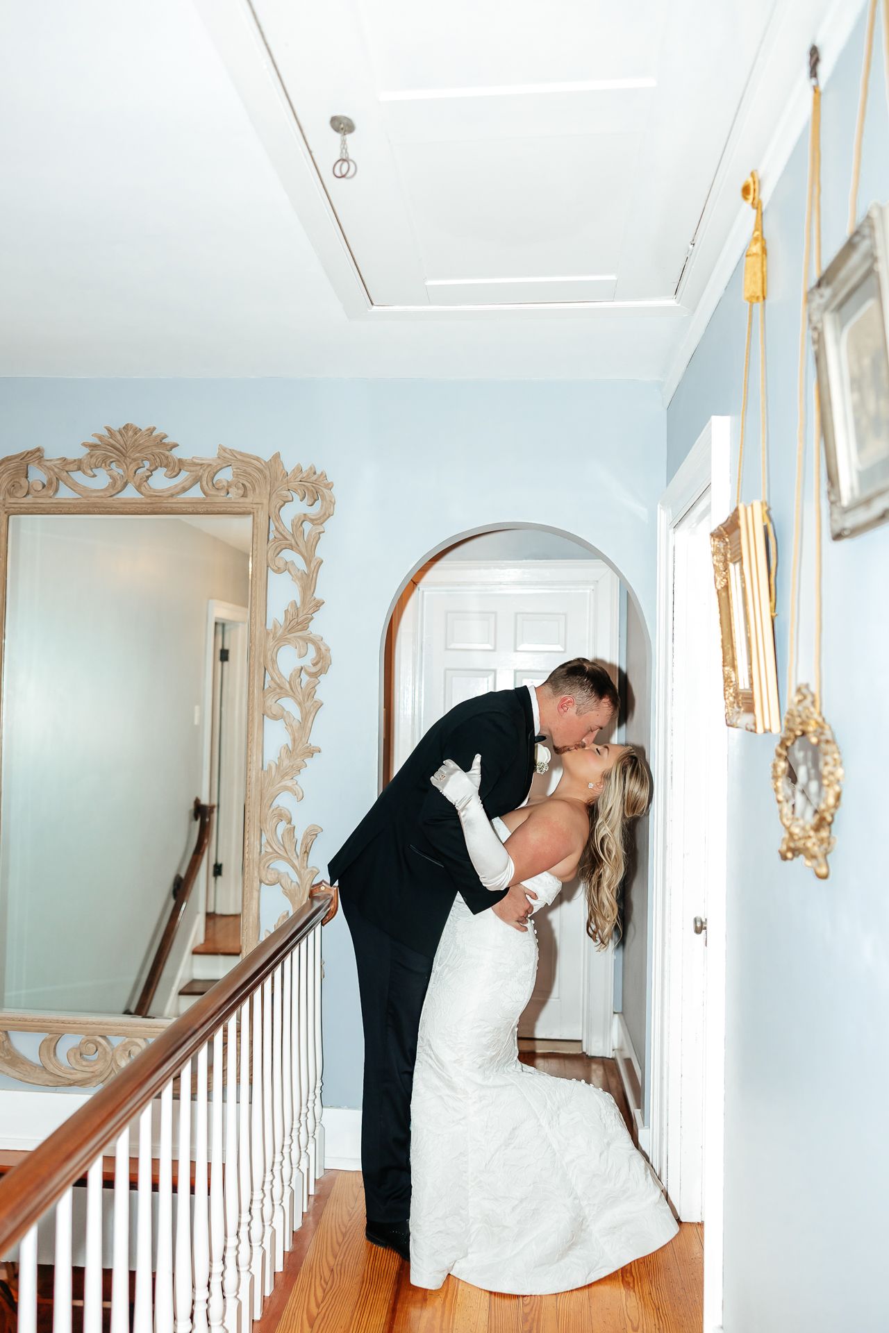 Groom kisses bride in hallway. Bride wears a white dress. Light blue walls, gold accents.