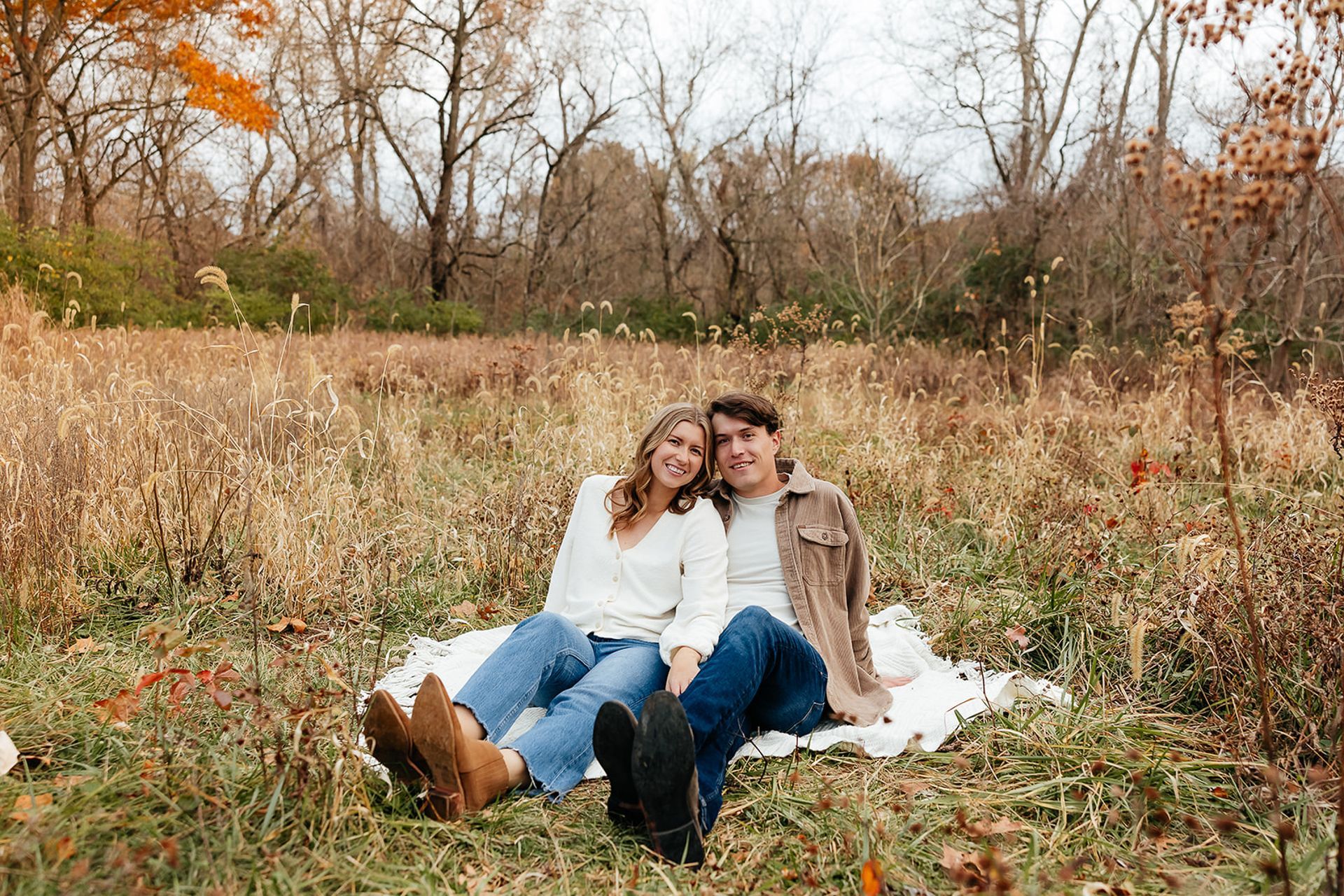 Couple sits on blanket in field, smiles, autumn foliage in background.