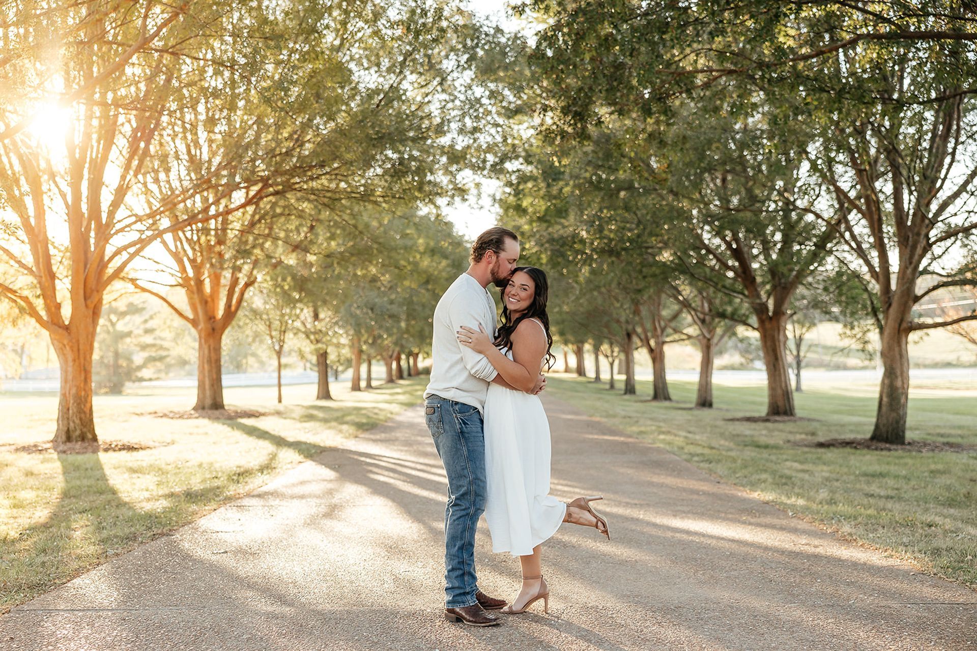 Couple embracing on a tree-lined path, sunlight streaming through trees. Man kissing woman’s head; both smiling.
