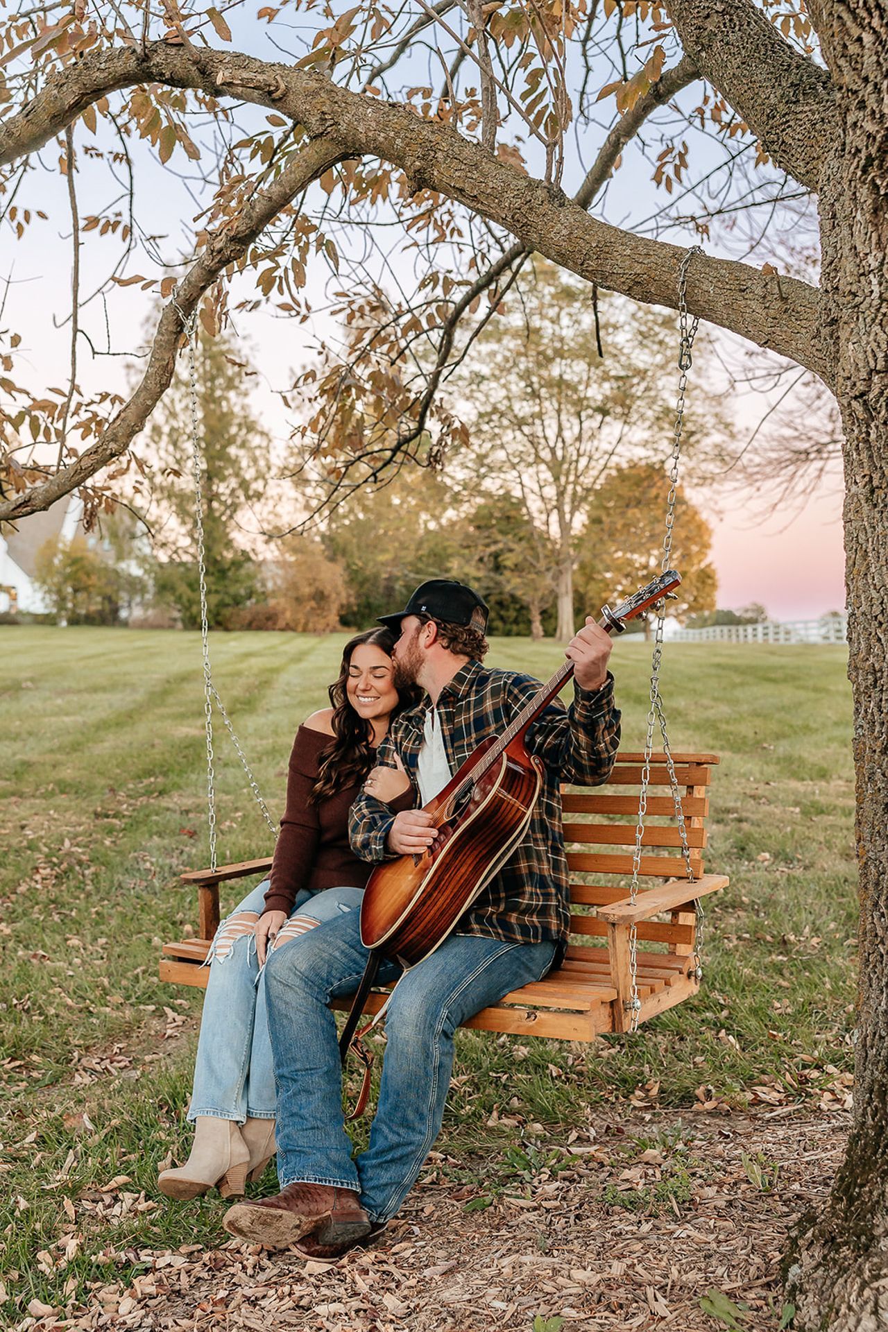 Couple on a wooden swing, the man playing guitar. Outdoors, trees, grass, and a house in the background.