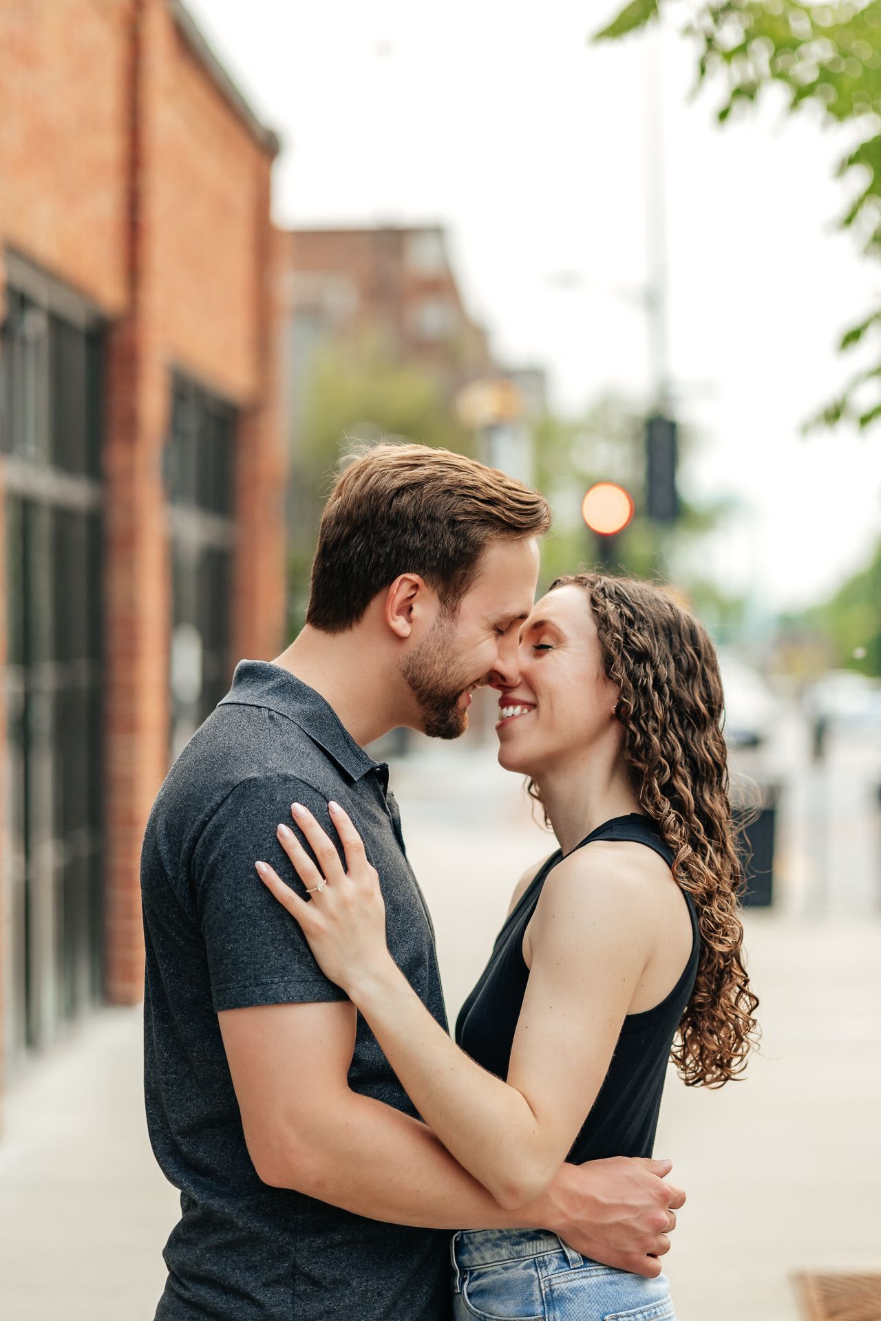 Couple embracing and smiling closely on a city sidewalk; brick building in background.