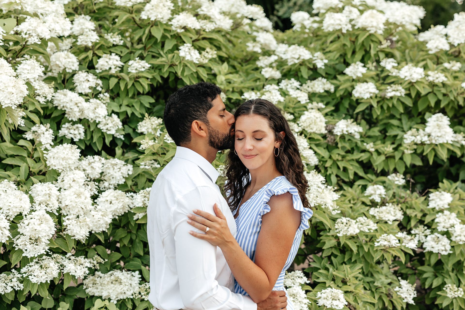 Couple embraces, man kisses woman's forehead, posing in front of white flowers.