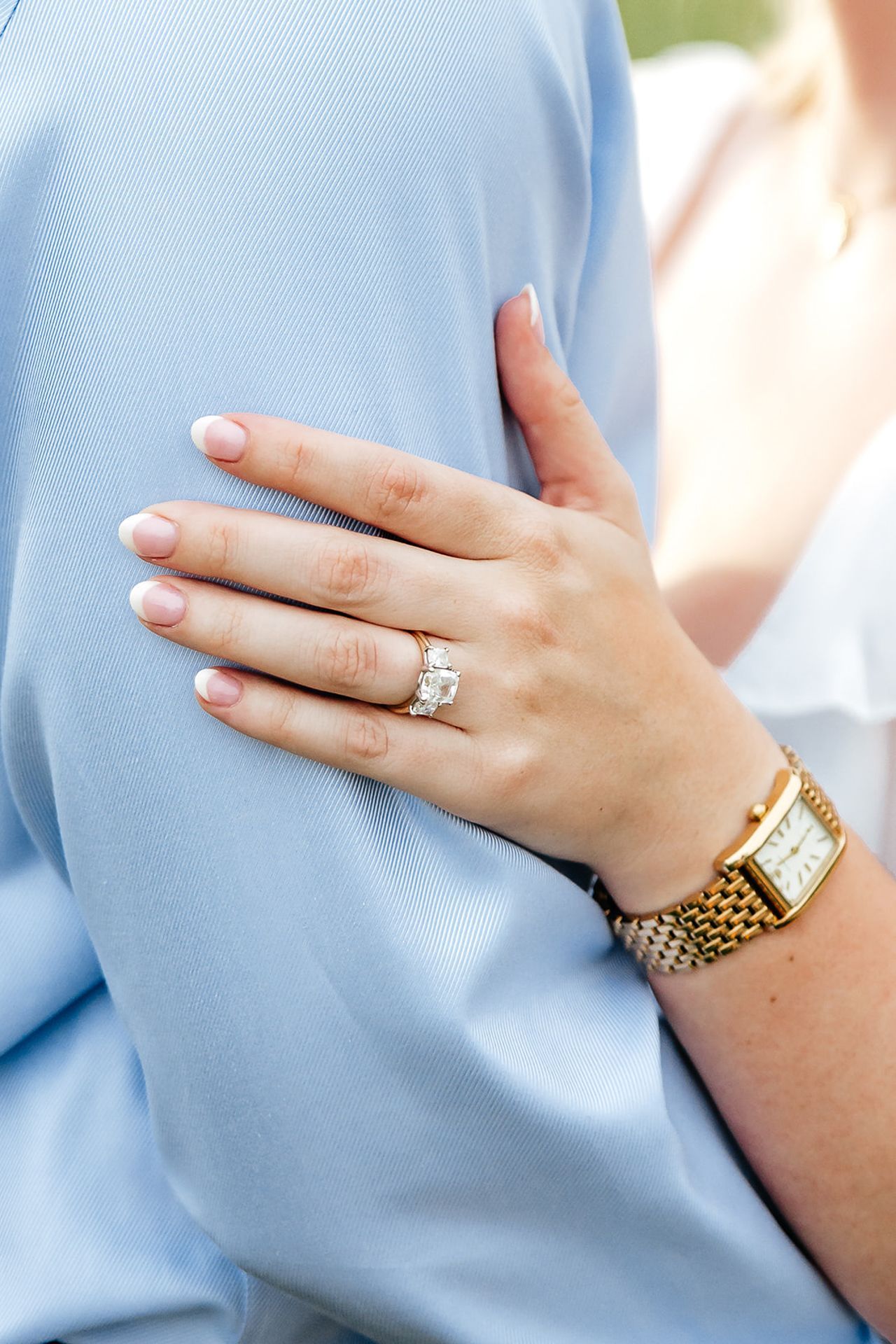 Woman's hand with diamond ring on man's arm. She wears a gold watch. Light blue shirt visible.