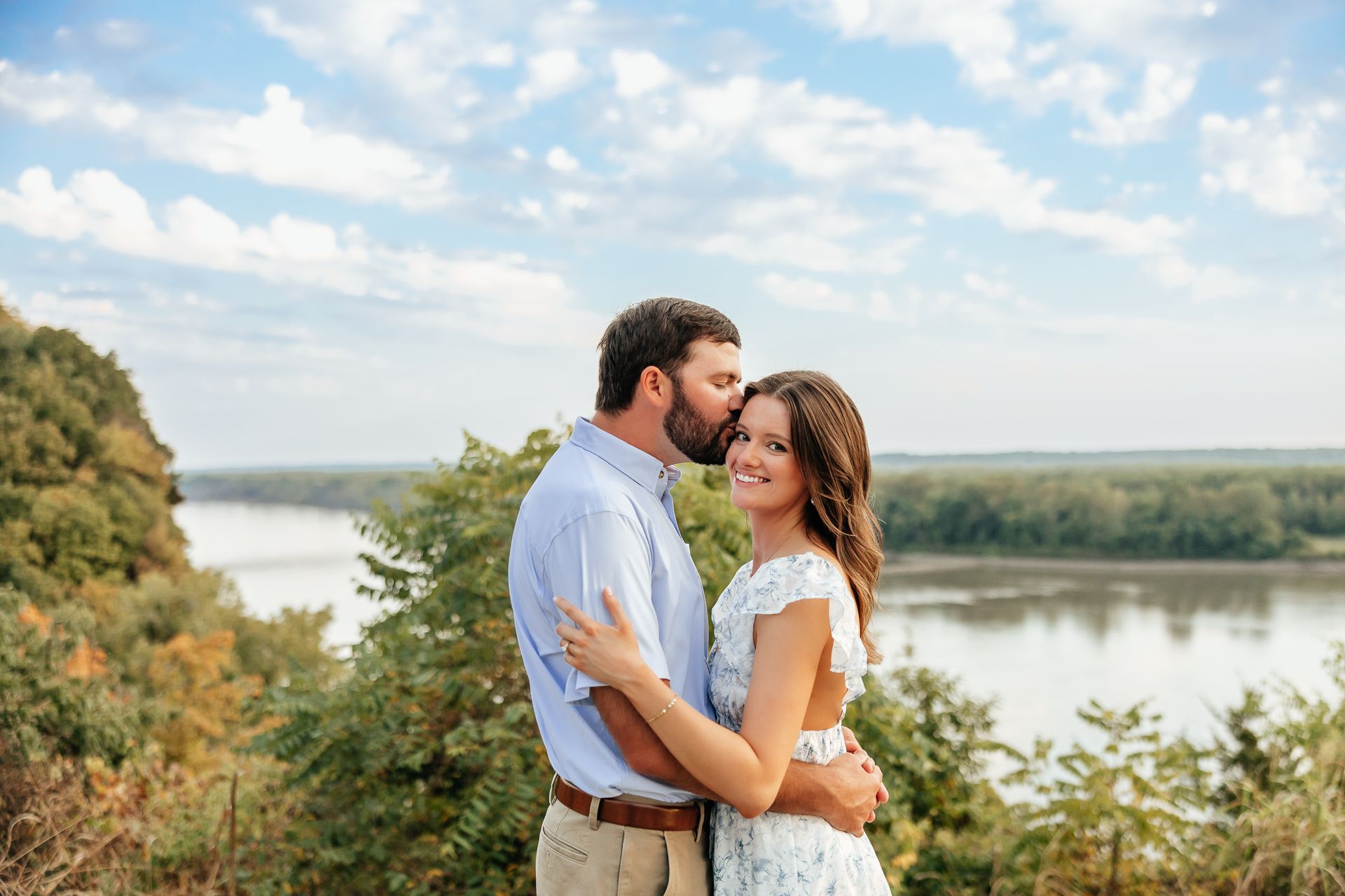 Couple embracing outdoors by a river, man kissing woman's forehead, blue sky background.