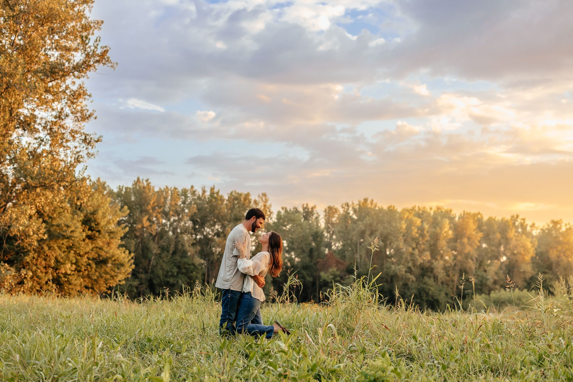 Couple embracing in a field during sunset, kissing; trees and golden light visible.