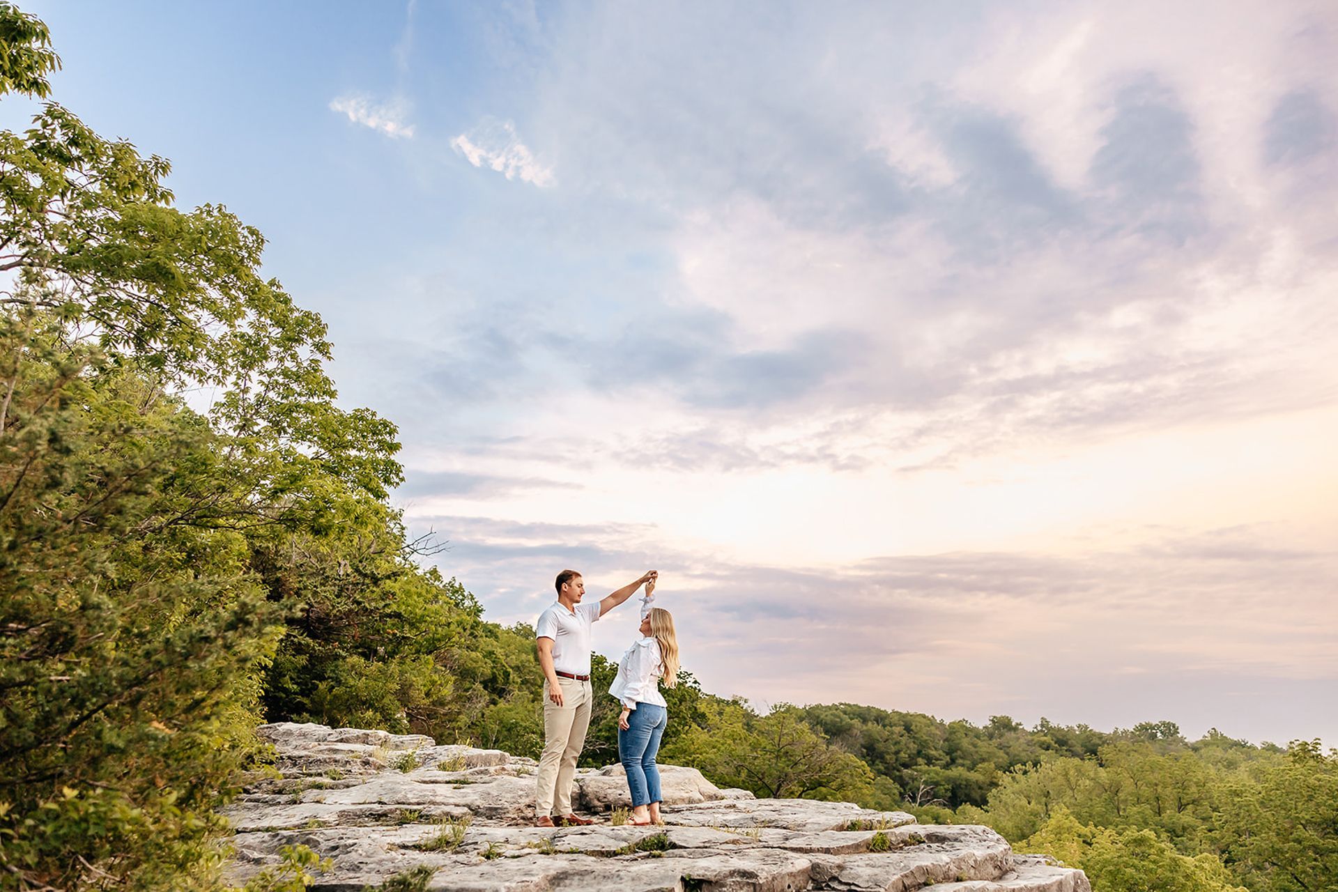 Couple dancing on a rocky outcrop overlooking a forested landscape, under a cloudy sky.