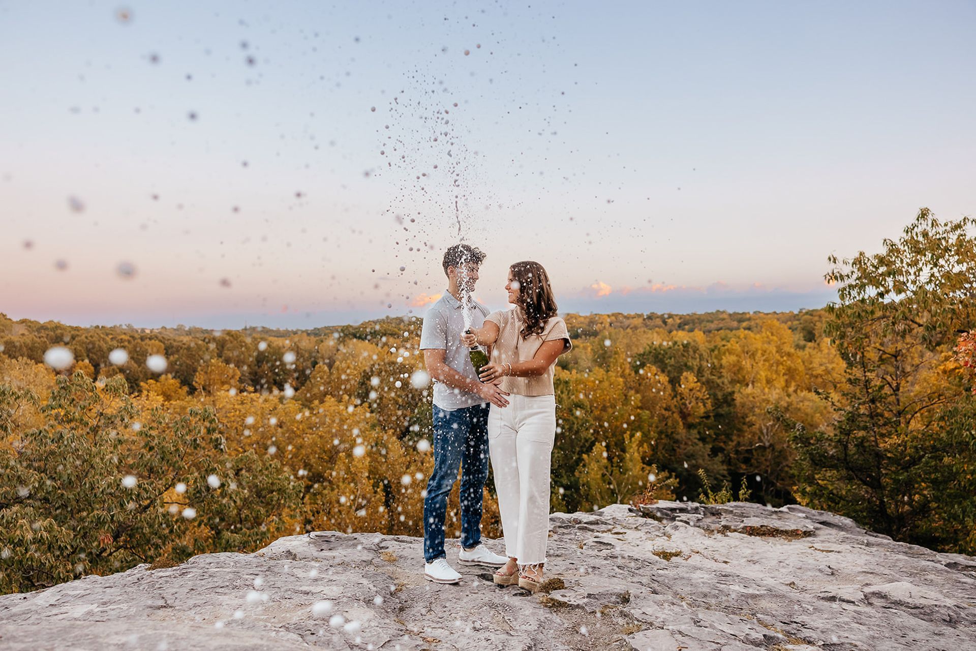 Couple on a cliff, spraying champagne. Trees with fall foliage in the background.