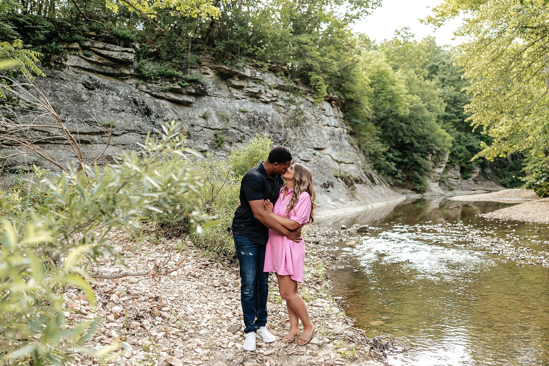 Couple kissing near a creek with rocky banks and trees in a natural setting. He wears black, she wears pink.
