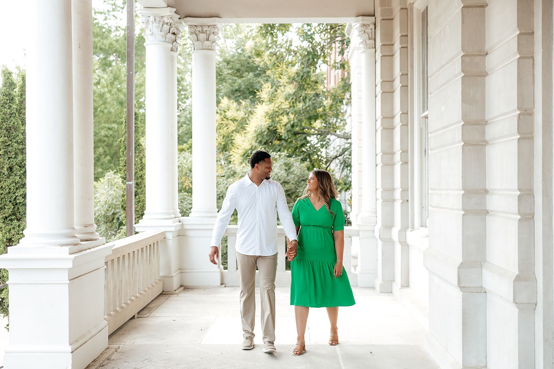 Couple holding hands, walking on a porch. The woman wears a green dress, the man a white shirt and khaki pants.
