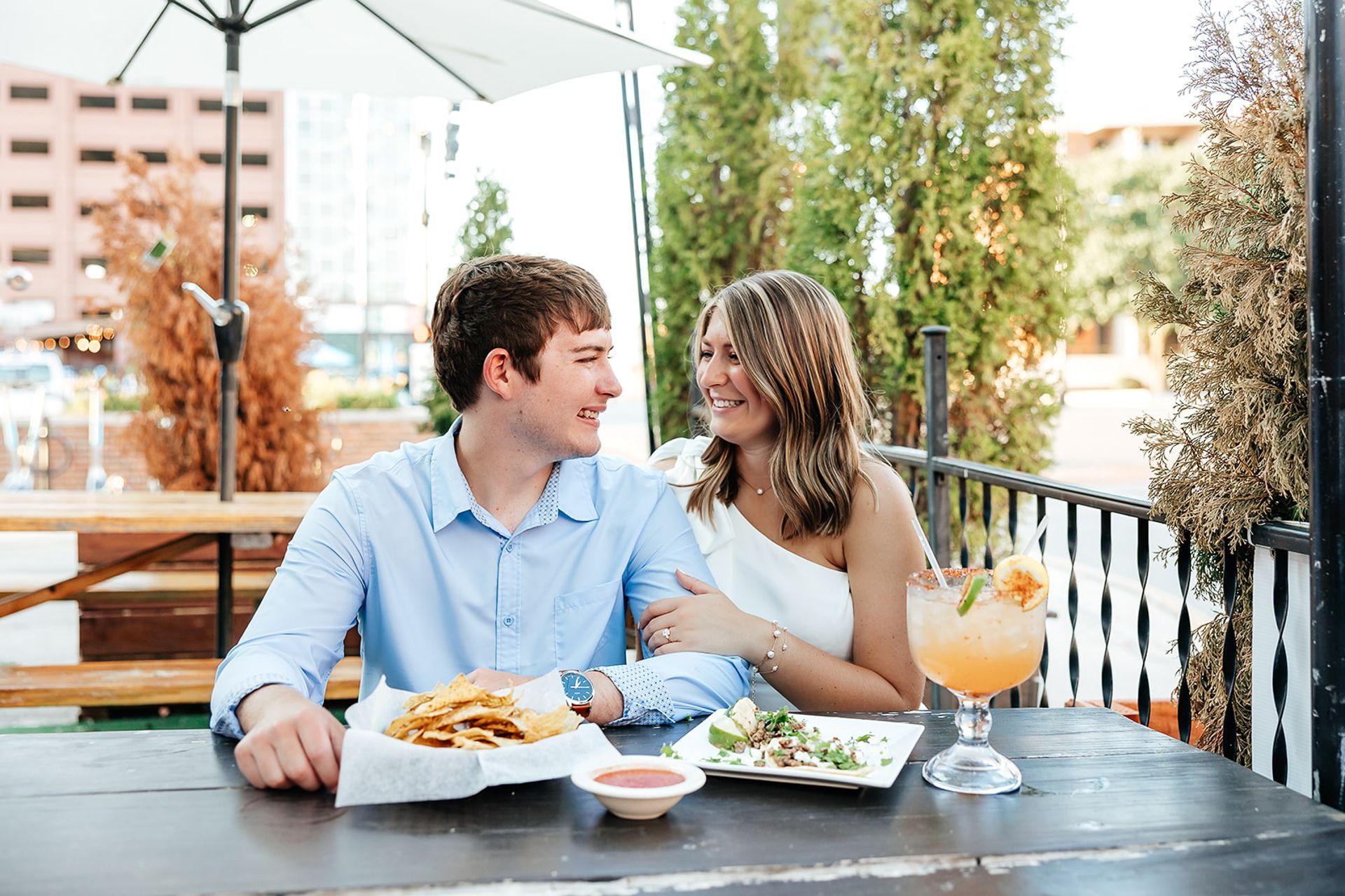 Couple smiles at each other over food and drinks at an outdoor restaurant.