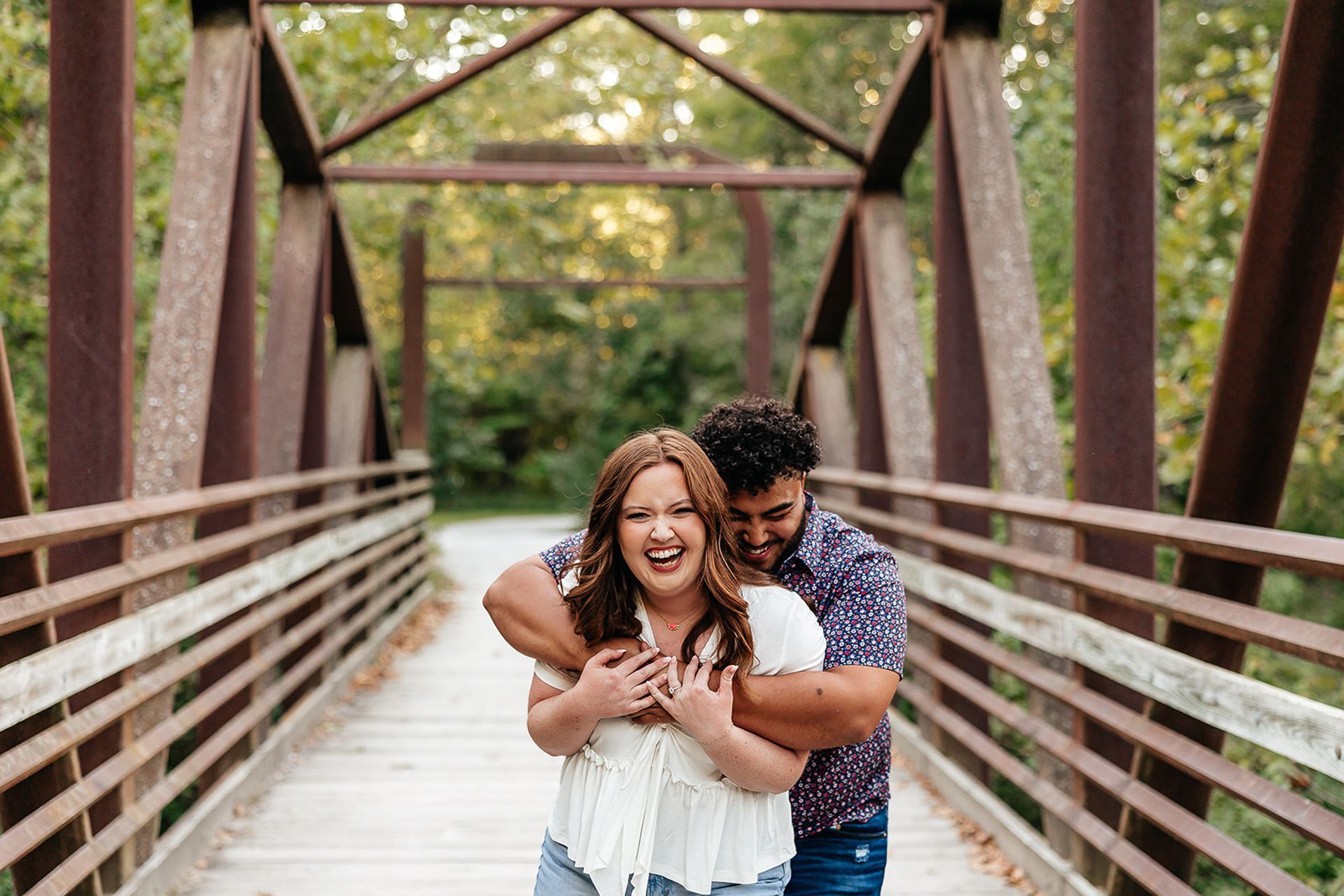 Couple hugging on a brown bridge, woman laughing, man smiling; trees in background.