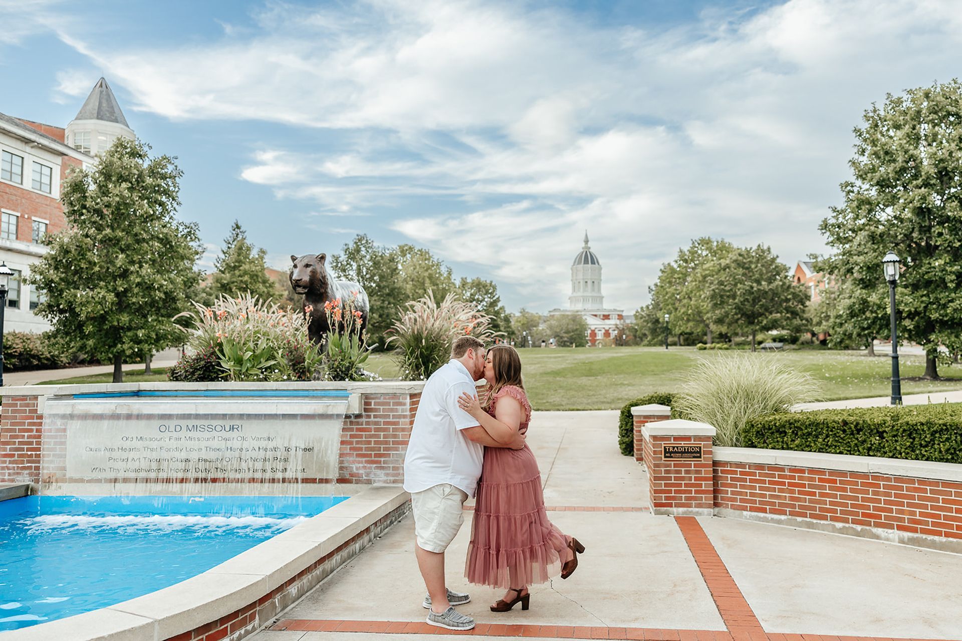 Couple kissing near a fountain on a college campus with a statue and buildings.