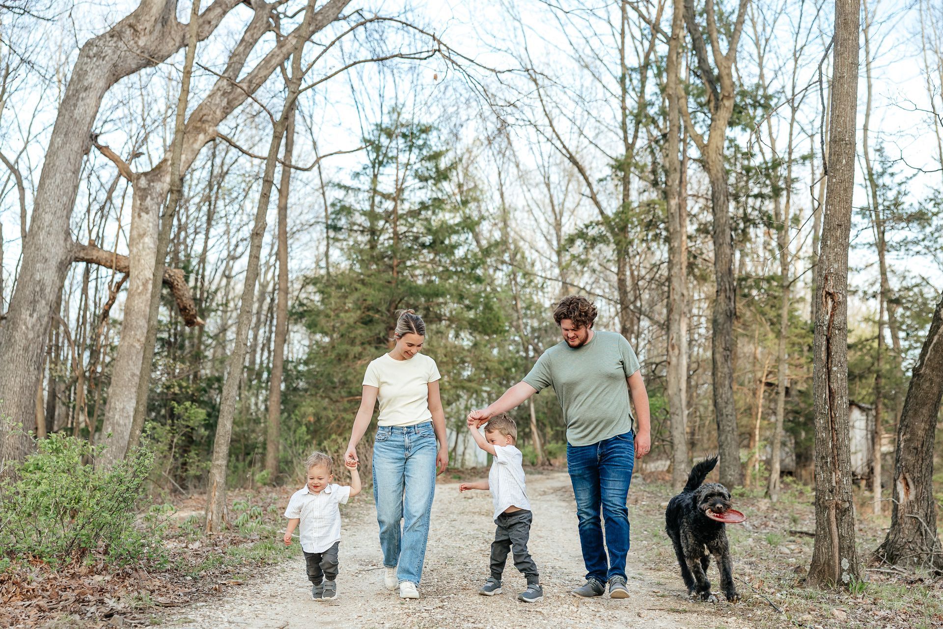 Family walks on a wooded path, holding hands with two children and a dog.