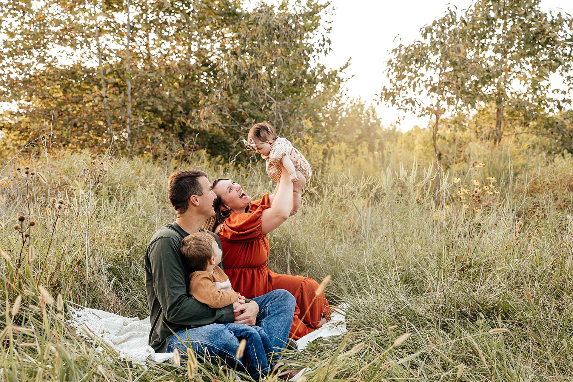 Family of four in a field; parents holding and playing with two small children, sitting on blanket in tall grass.