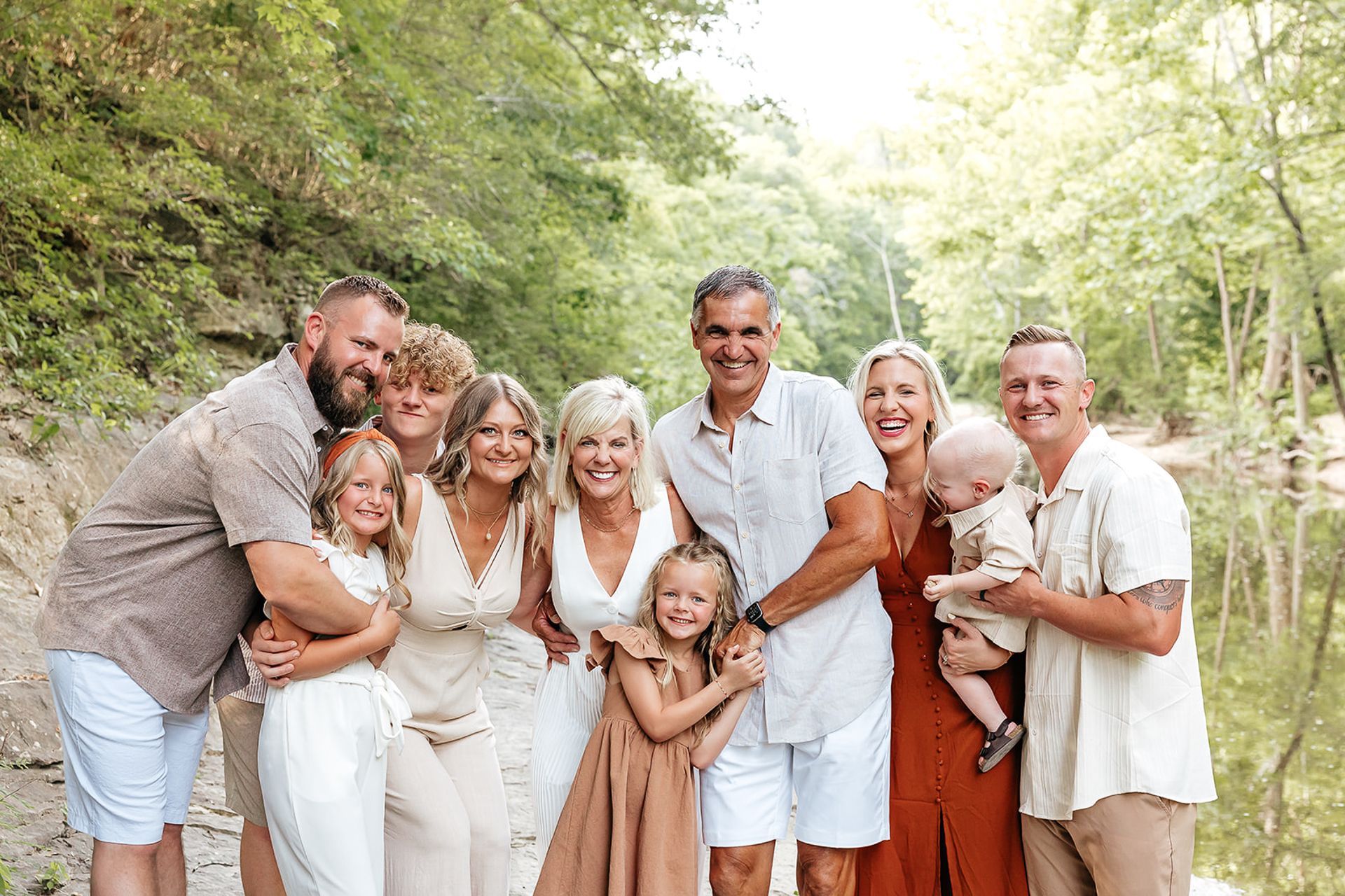 Family group posing by a creek. People in neutral-toned clothing smiling. Lush green foliage surrounds.