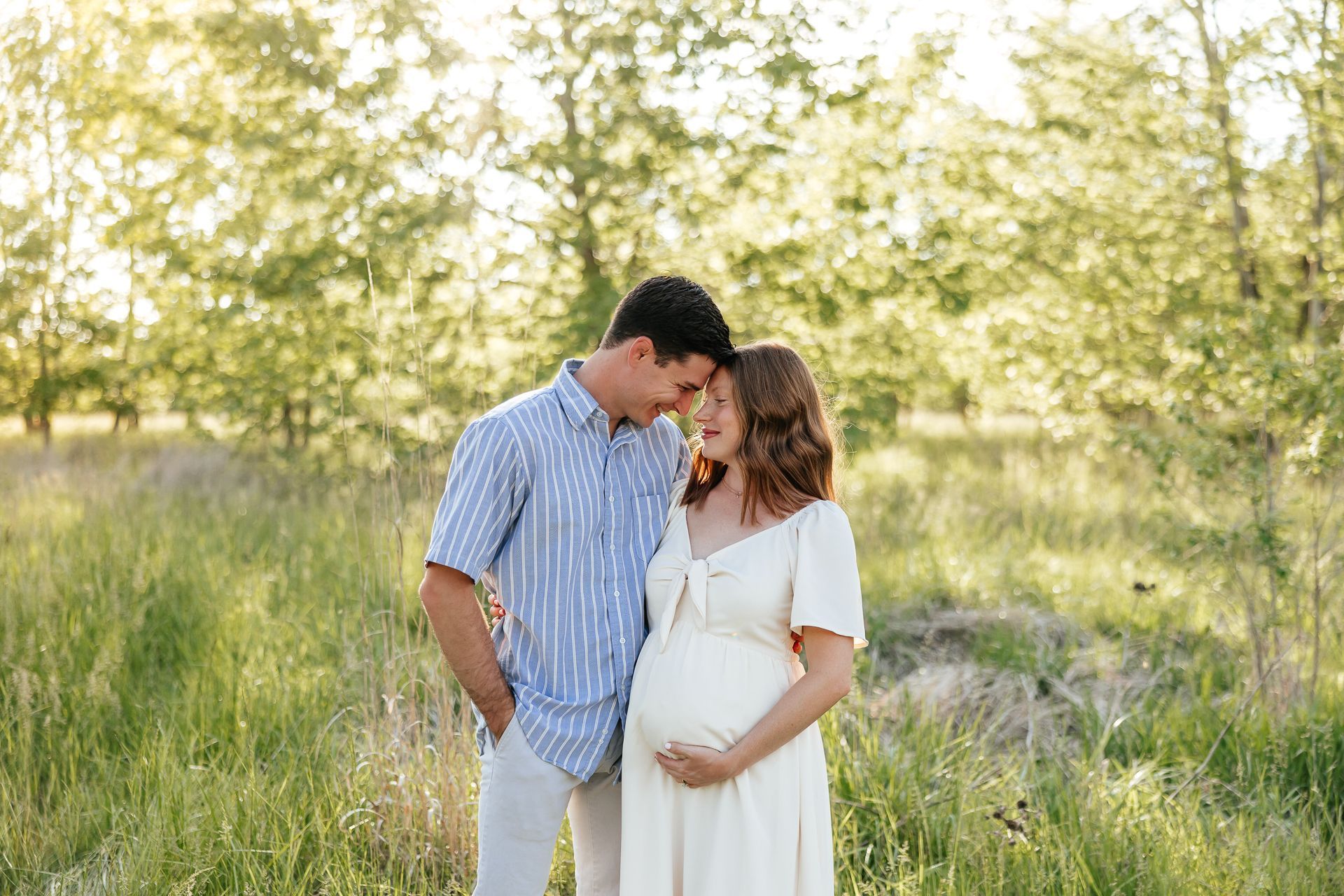 Pregnant woman and man embrace outdoors, golden light. Woman holds belly, man looks at her.